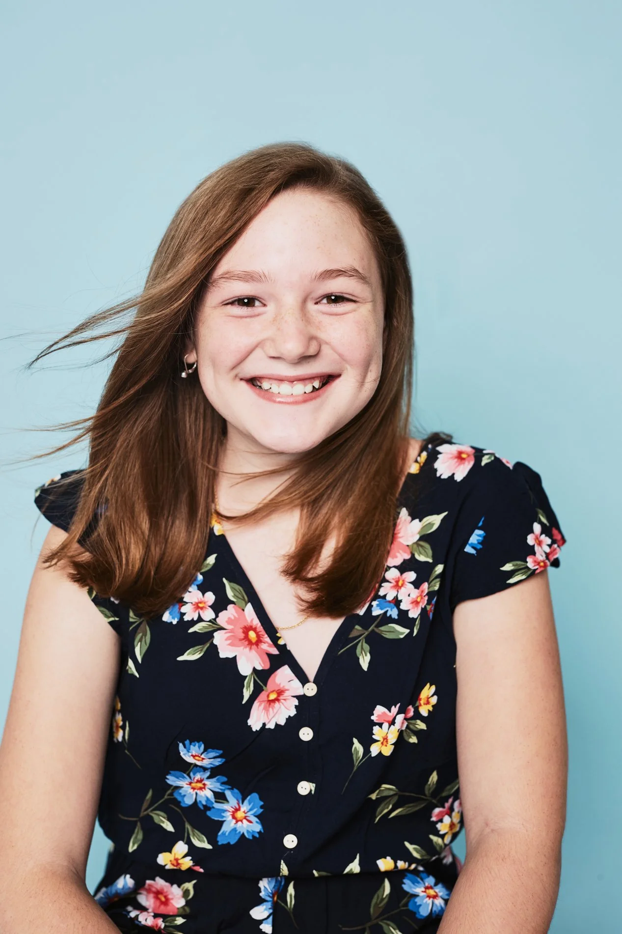 A young girl with brown hair, wearing a navy blue floral dress, smiling against a light blue background.
