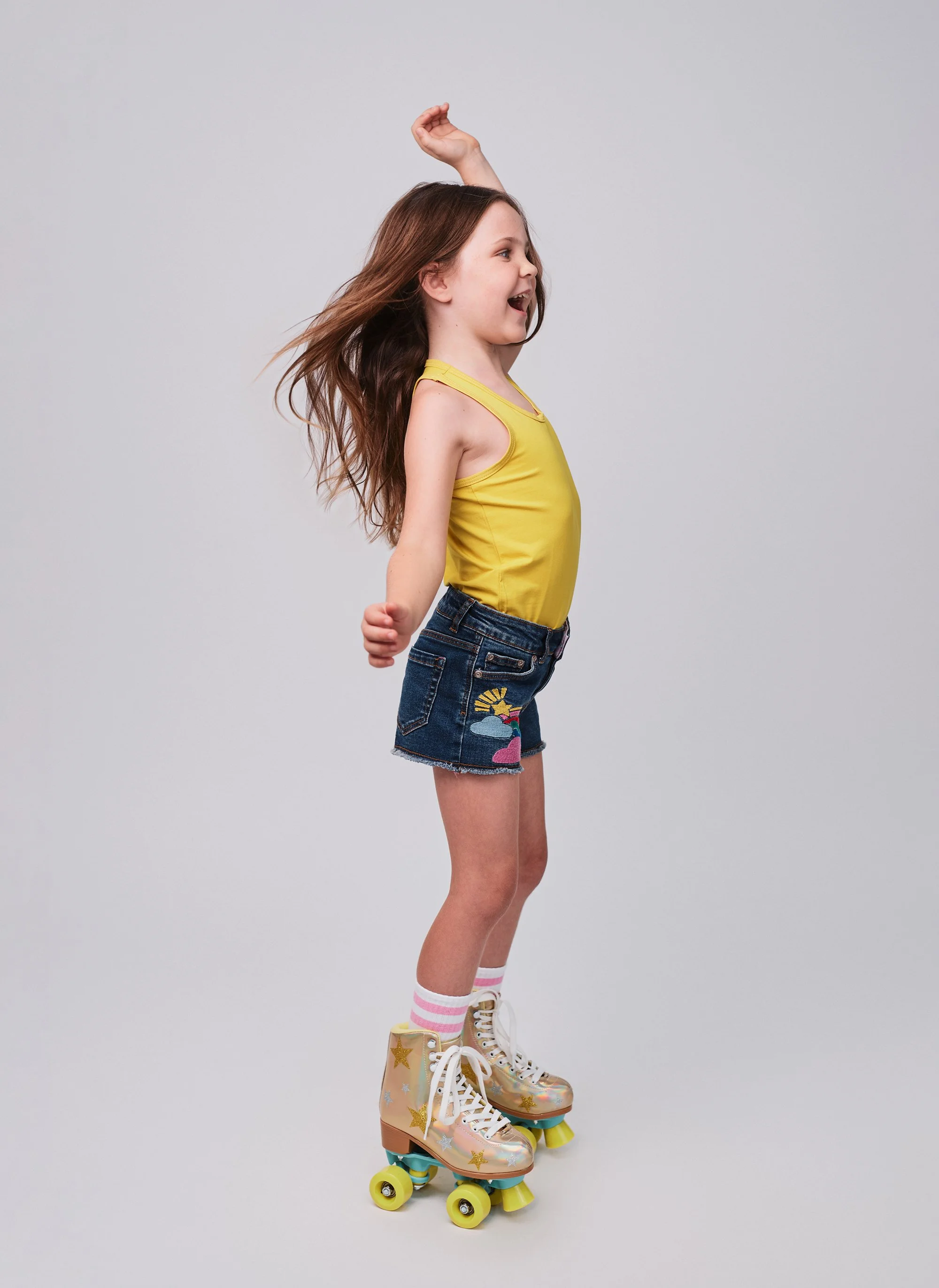 A young girl wearing a yellow top, denim shorts with a unicorn patch, and pink and white striped socks, stands on roller skates with a rainbow-colored sole, on a plain gray background. She appears joyful and energetic.