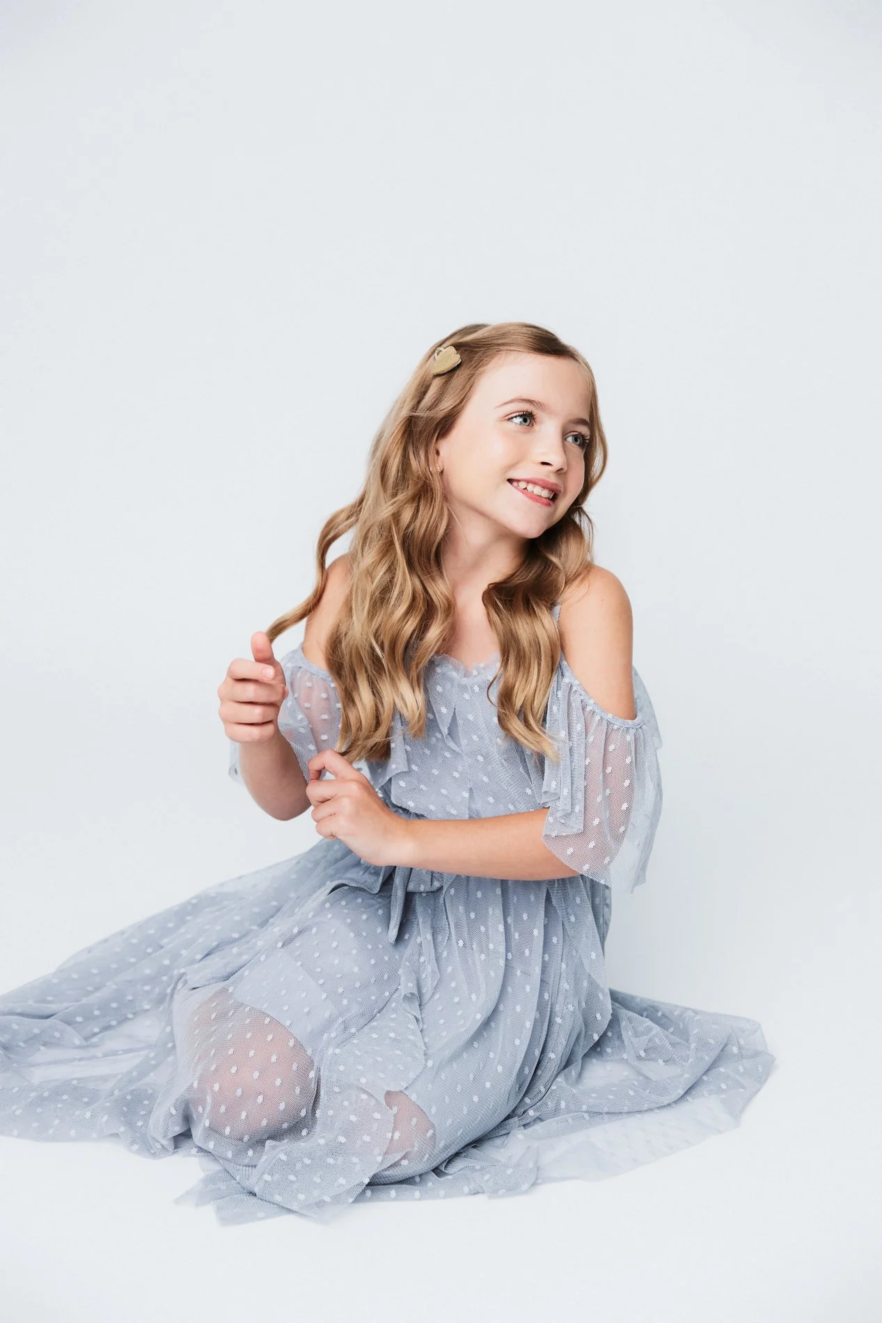 Young girl with long wavy blonde hair sitting on the floor, wearing a light blue polka dot dress, smiling and looking to the side, against a plain white background.
