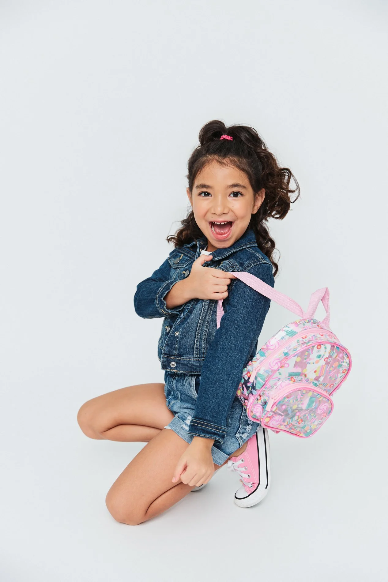 A young girl with brown curly hair wearing a denim jacket, denim shorts, pink sneakers, and a pink backpack with colorful patterns.