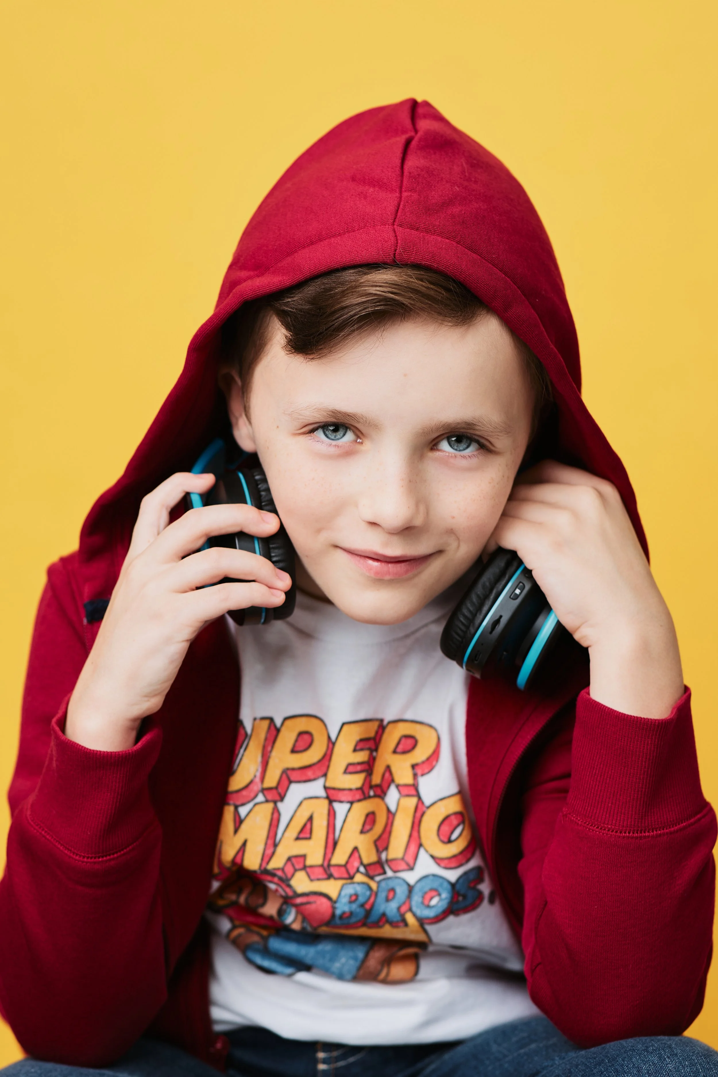 A young boy with blue eyes, wearing a red hoodie and a 'Super Mario Bros.' T-shirt, listening to headphones against a yellow background.