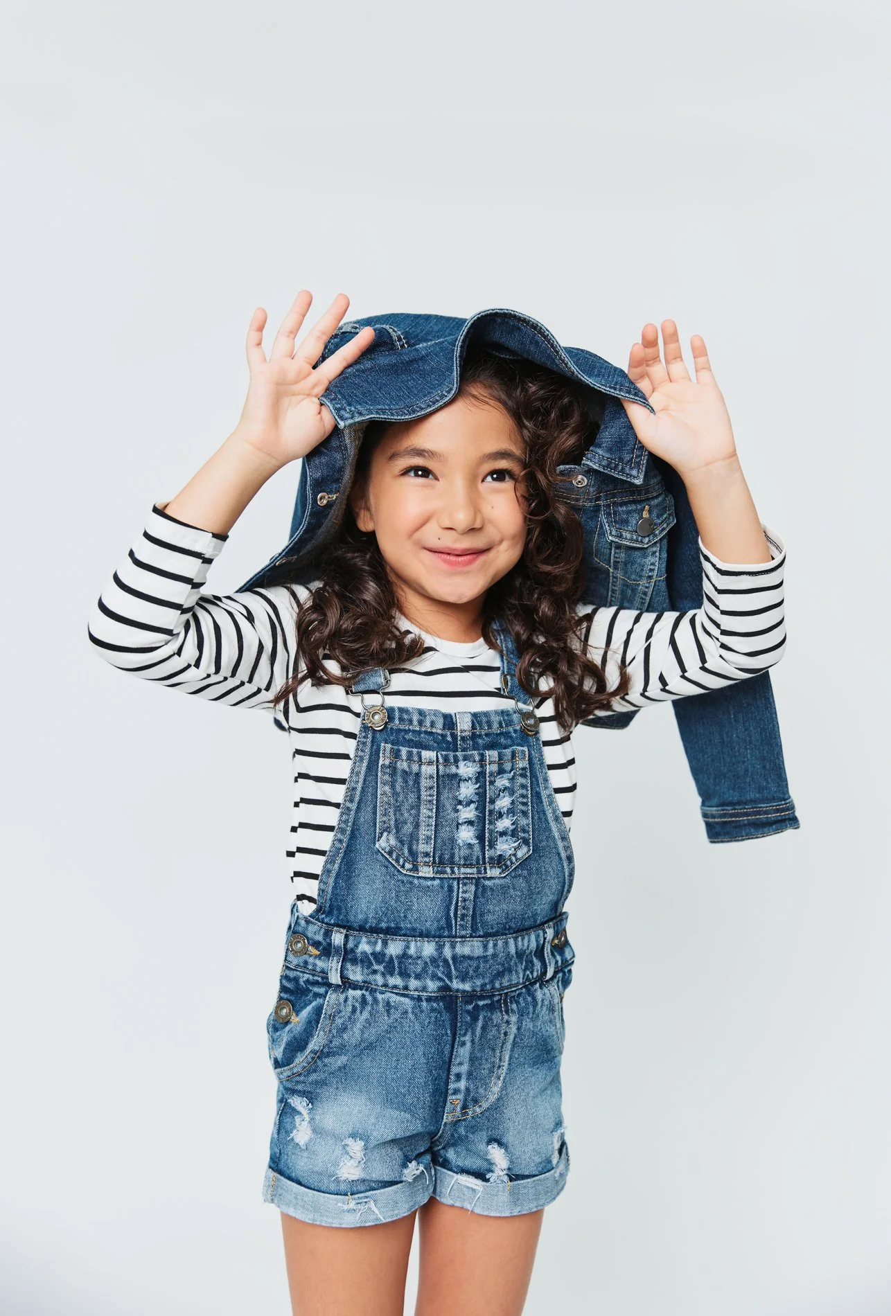 Young girl with curly hair smiling, wearing a striped shirt and denim overalls, holding a denim jacket over her head.