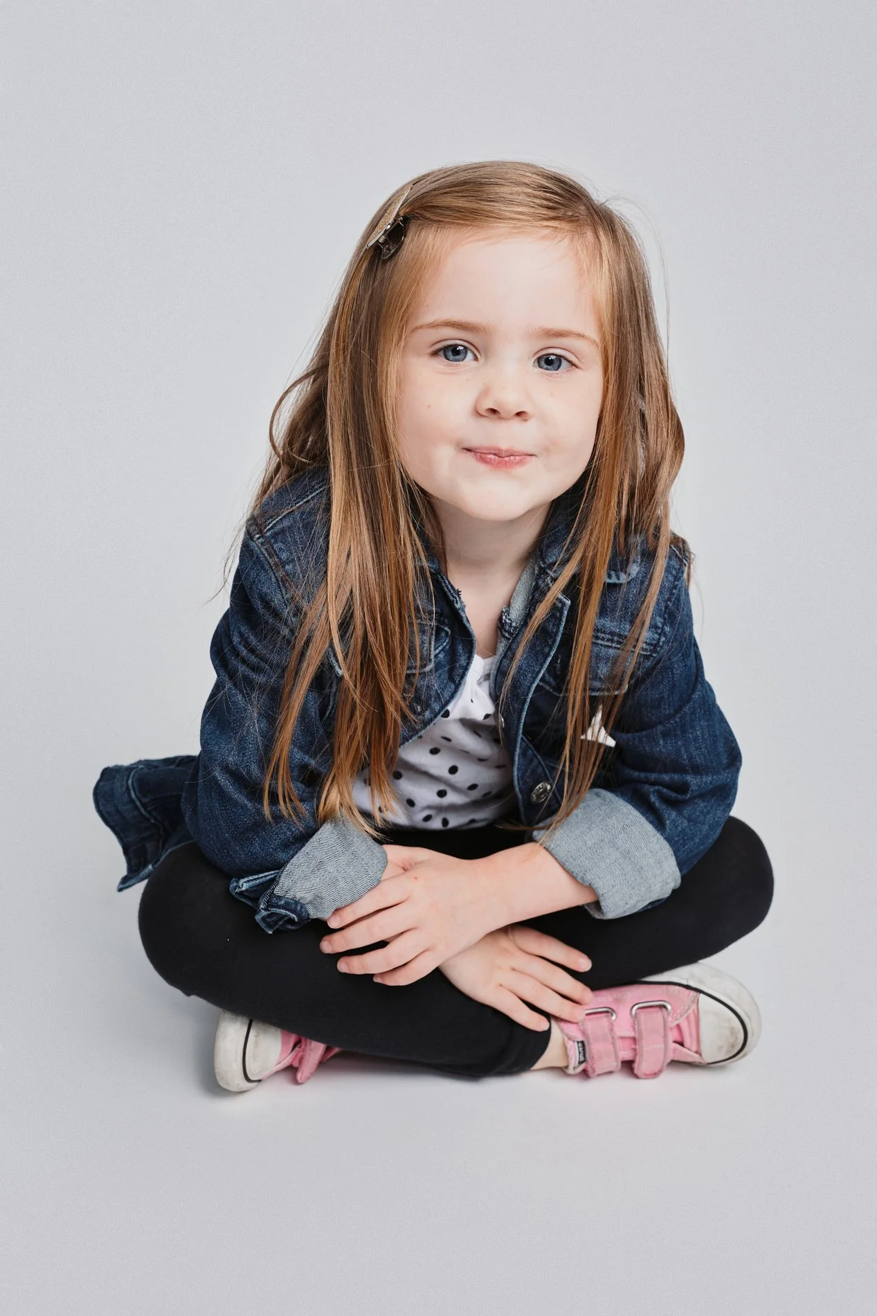 Houston Children's Photograper |A young girl with blonde hair, blue eyes, and freckles sitting cross-legged on the floor, wearing a denim jacket, polka dot shirt, black pants, and pink sneakers, with a hair clip on her hair
