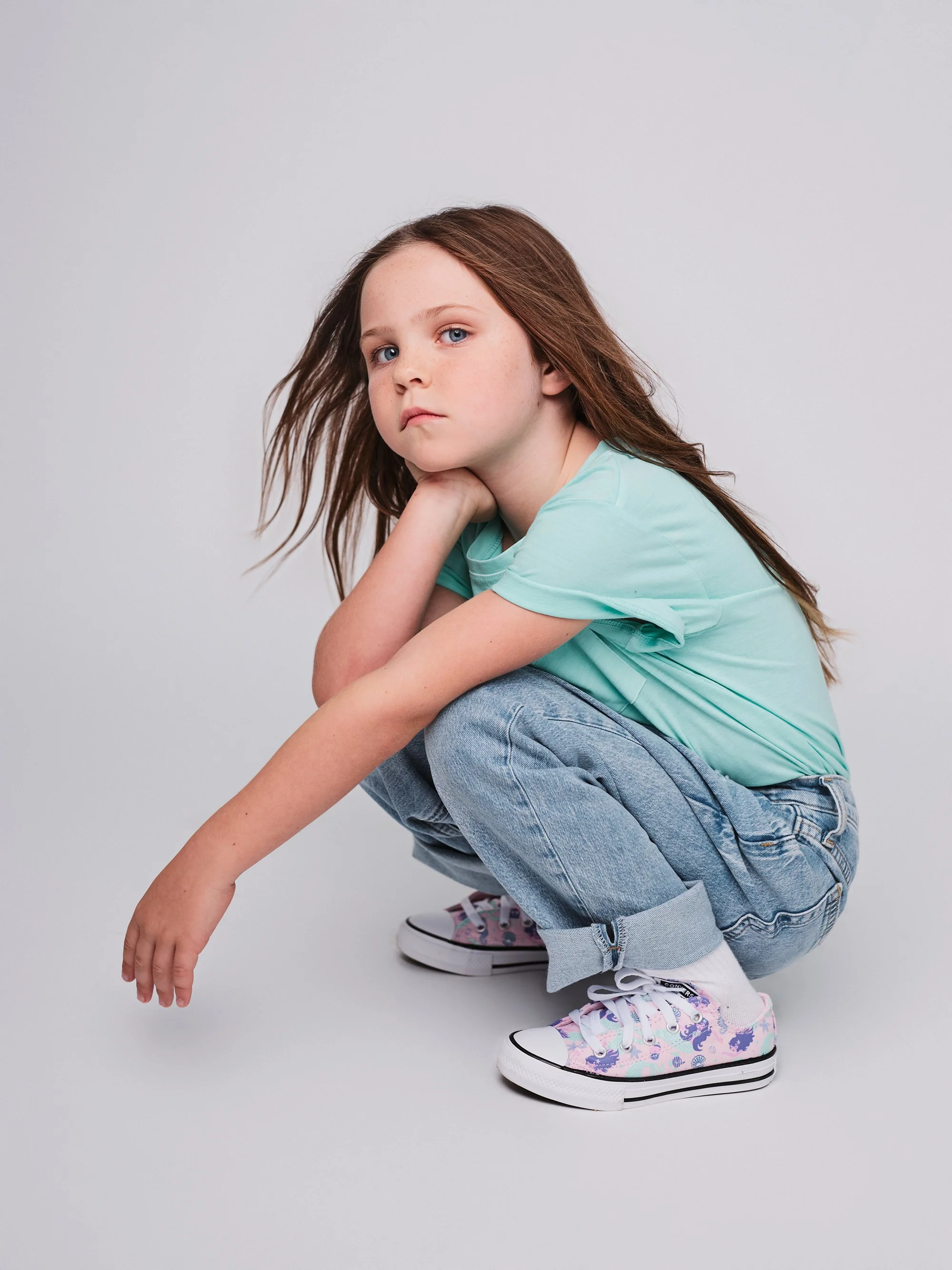 A young girl with long brown hair, blue eyes, and freckles, crouching down with one knee on the ground, resting her chin on her hand, wearing a light blue t-shirt, rolled-up jeans, and colorful sneakers against a plain white background.