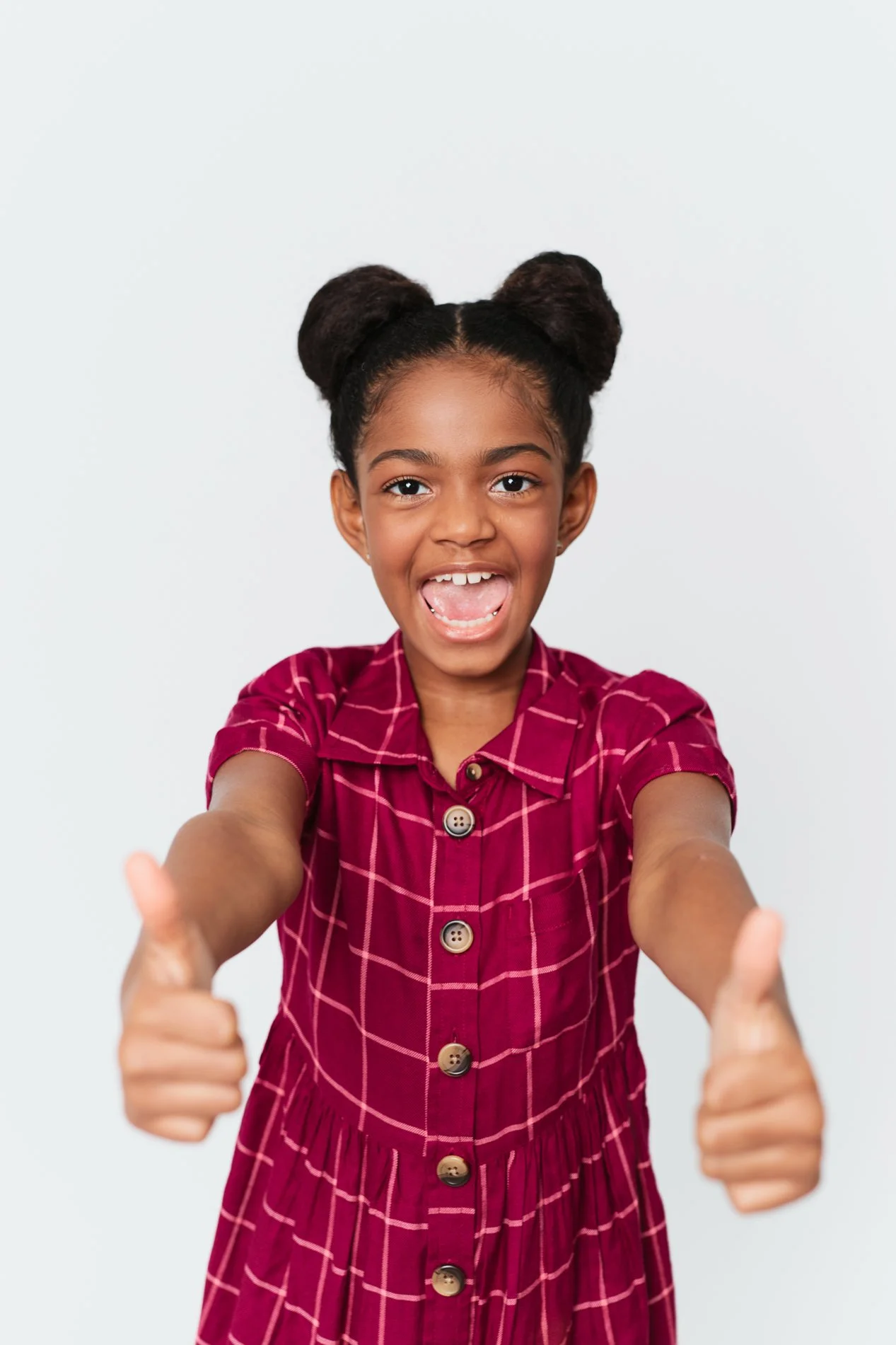 A young girl with dark hair styled in two buns, smiling and giving thumbs up, wearing a red checkered dress, reaching towards the camera with her arms extended.
