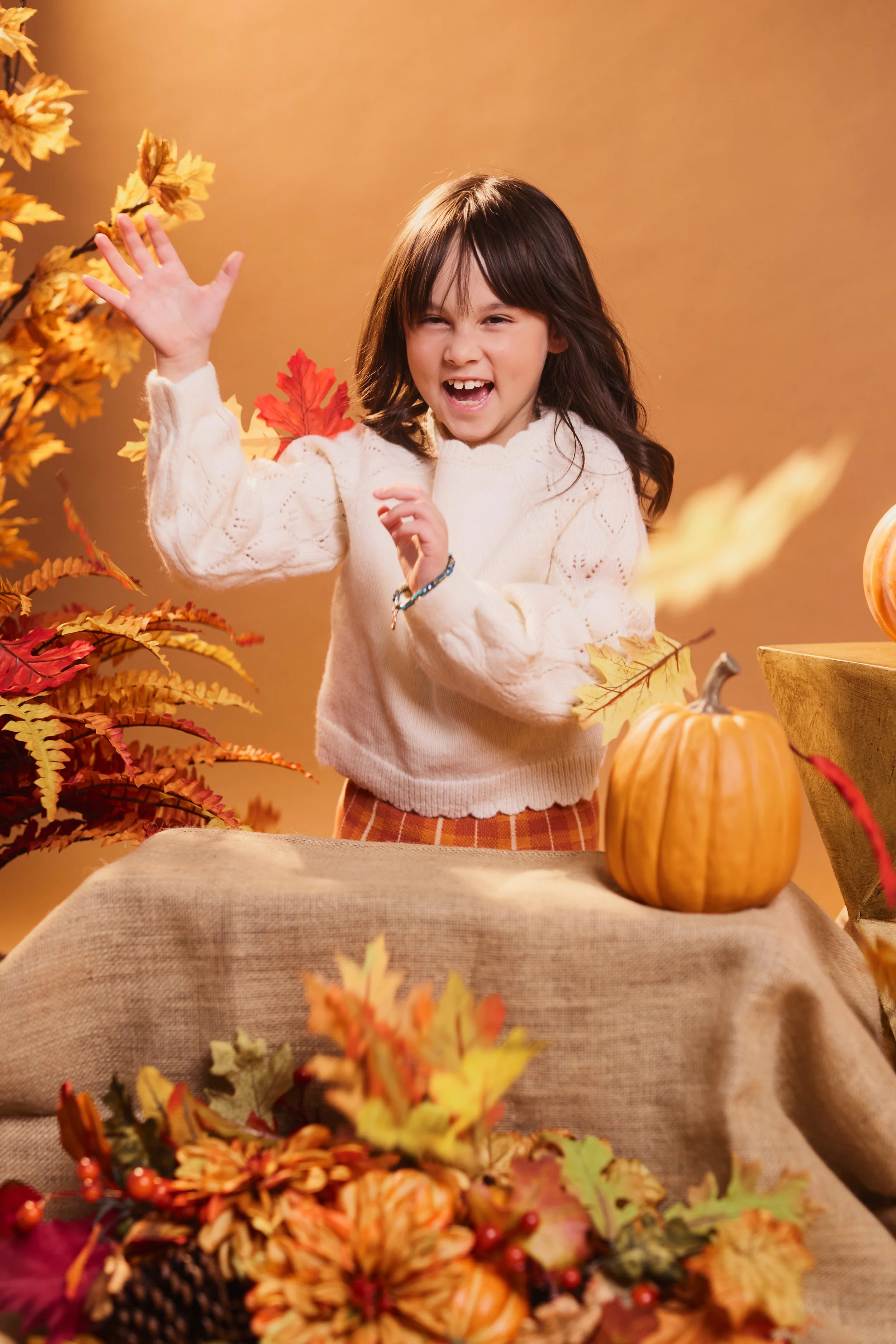 A young girl with dark hair, wearing a white sweater, is smiling and waving her hand. She is surrounded by autumnal decorations, including pumpkins and colorful fall leaves. The background is a warm orange tone.