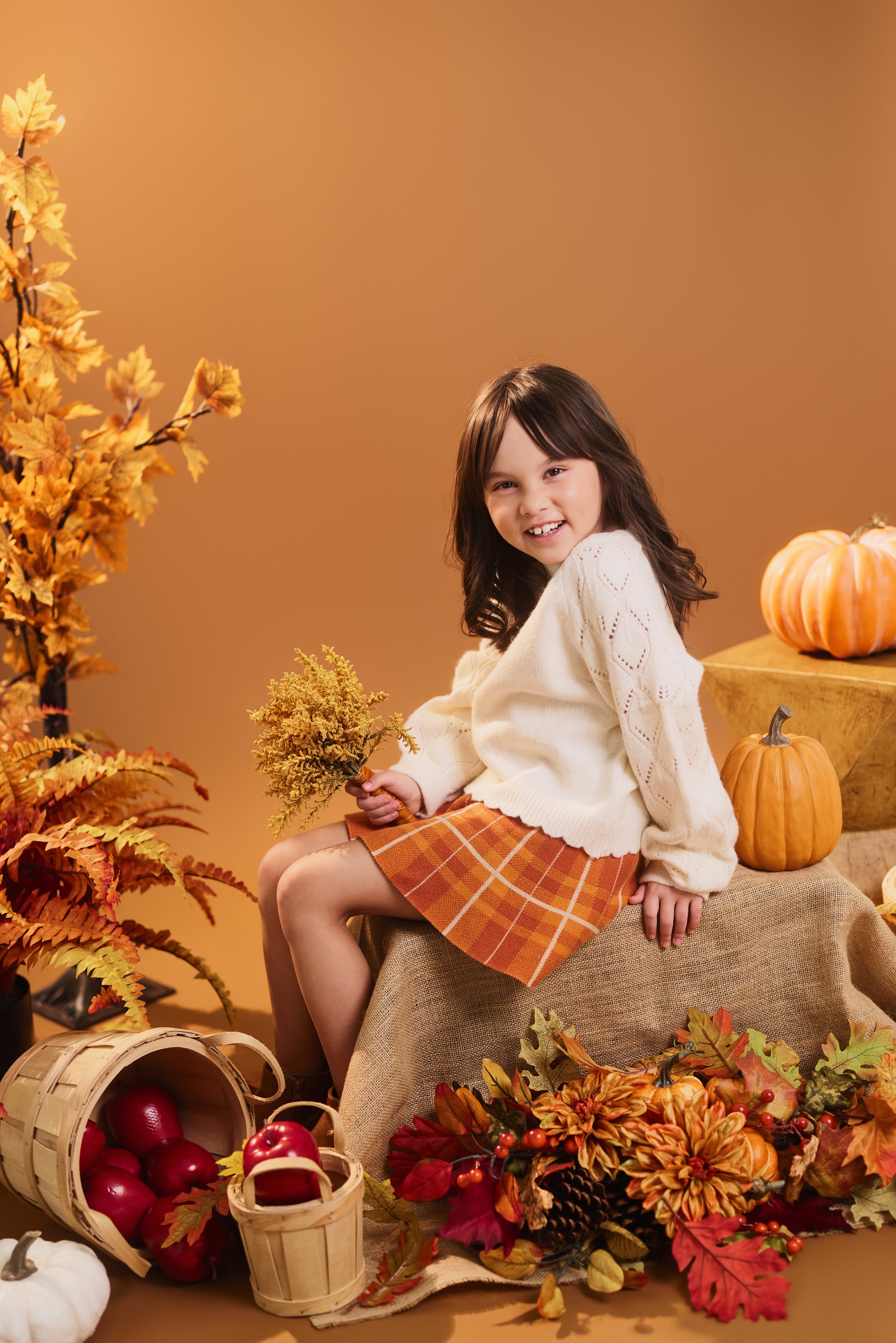 A young girl with dark hair and a beige sweater sitting among fall-themed decorations, pumpkins, apples, flowers, and leaves in warm autumn colors, against a light brown background.