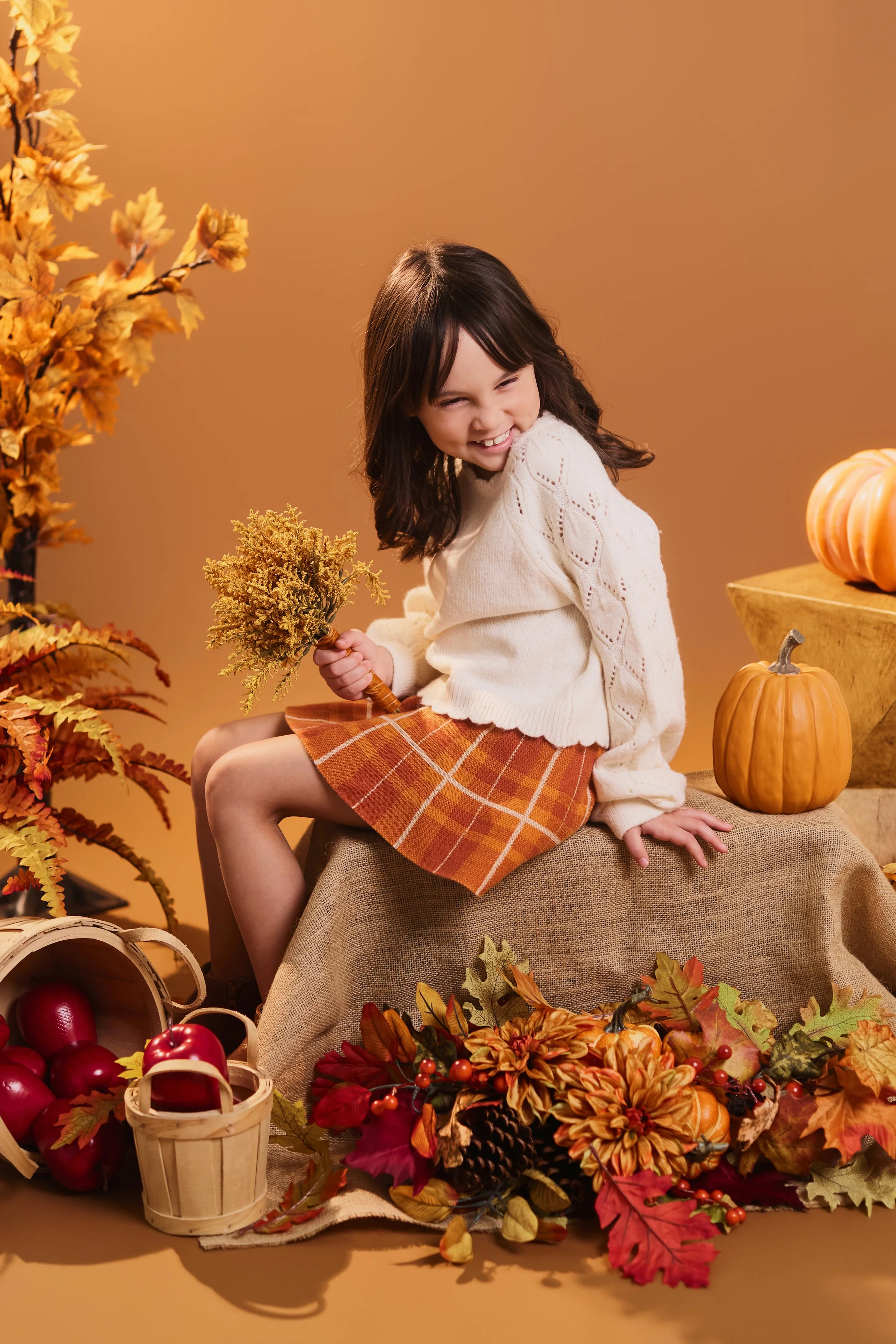 Young girl with dark hair, wearing a white sweater and orange plaid skirt, sitting on a table surrounded by autumnal decorations like pumpkins, apples, fall leaves, and yellow flowers, with a warm orange background.