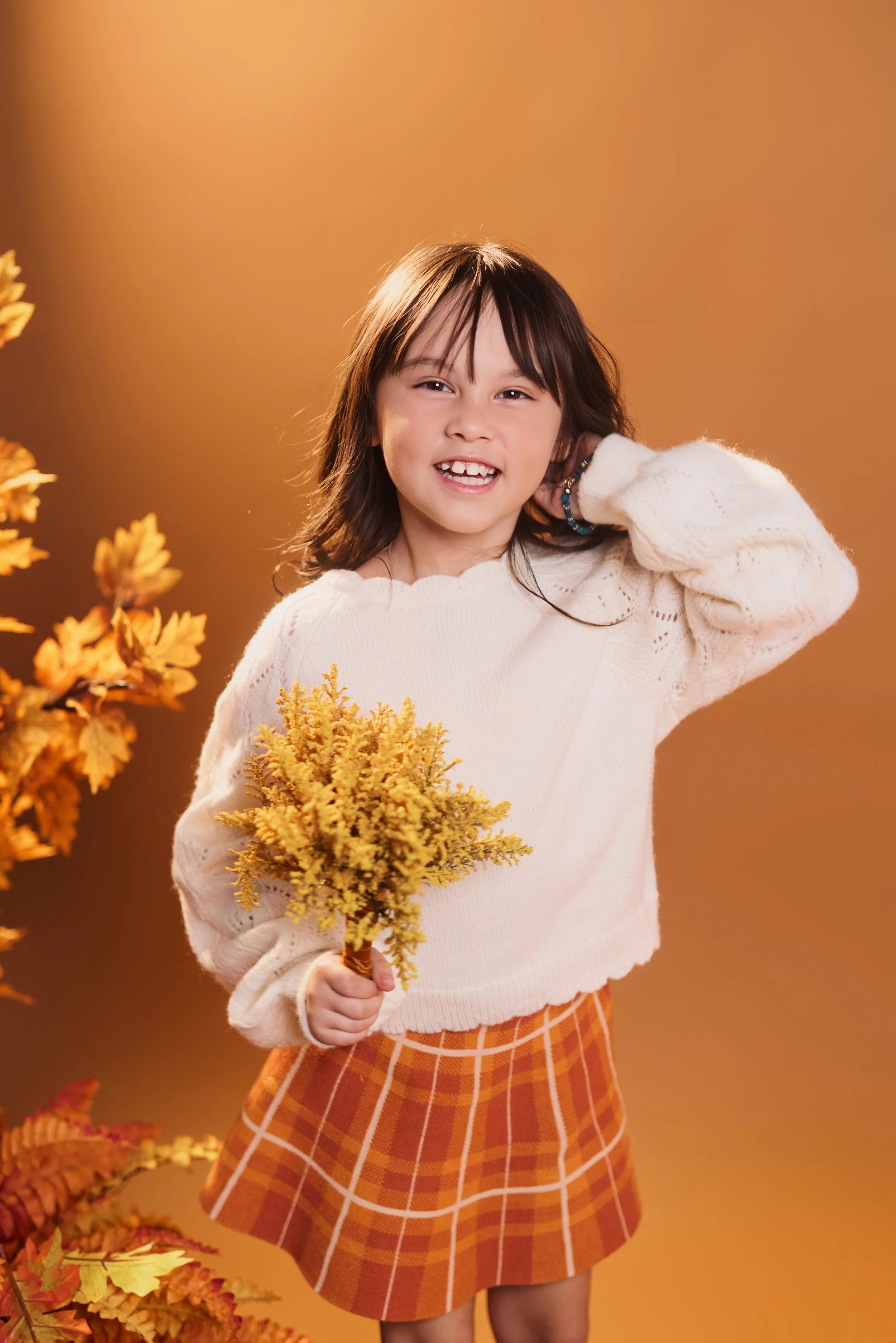 A young girl with brown hair smiling and holding a bunch of yellow flowers, standing against an orange background surrounded by autumn leaves.