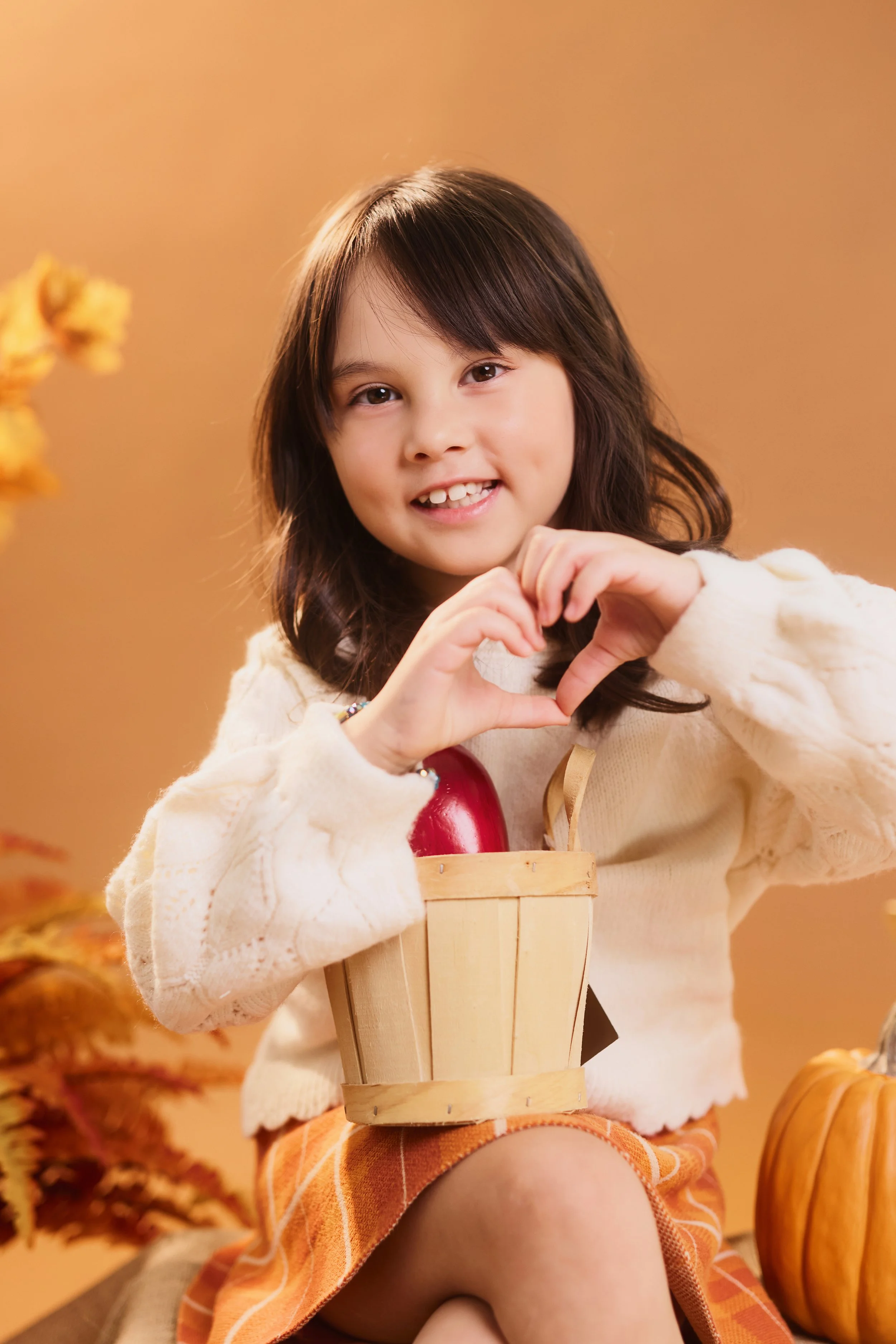 A young girl with brown hair smiling and making a heart shape with her hands, sitting with a basket and surrounded by pumpkins and autumn decor.