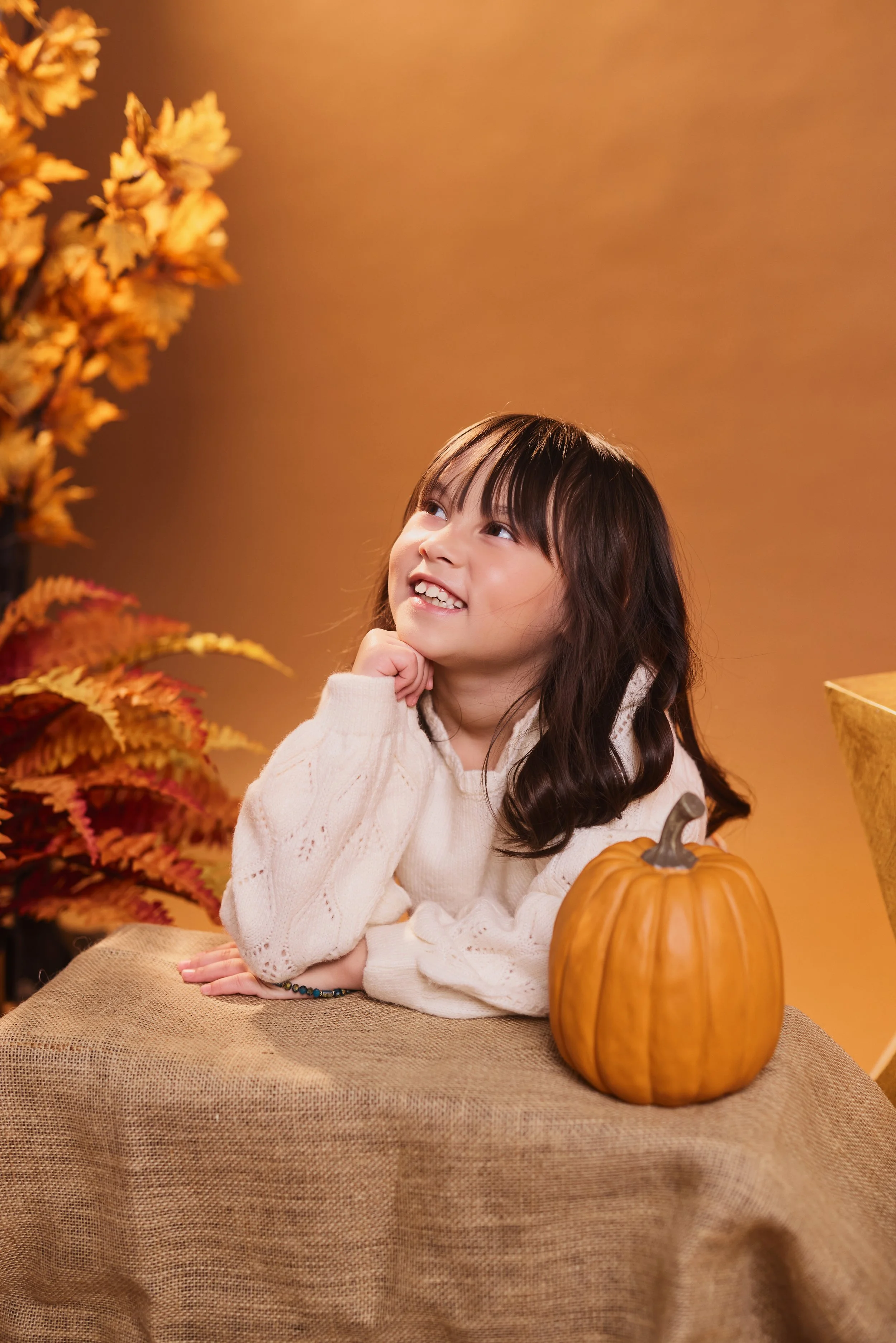 A young girl with long dark hair, wearing a beige sweater, sitting at a table with a pumpkin in front of her, surrounded by autumn-colored leaves and a warm, orange background.