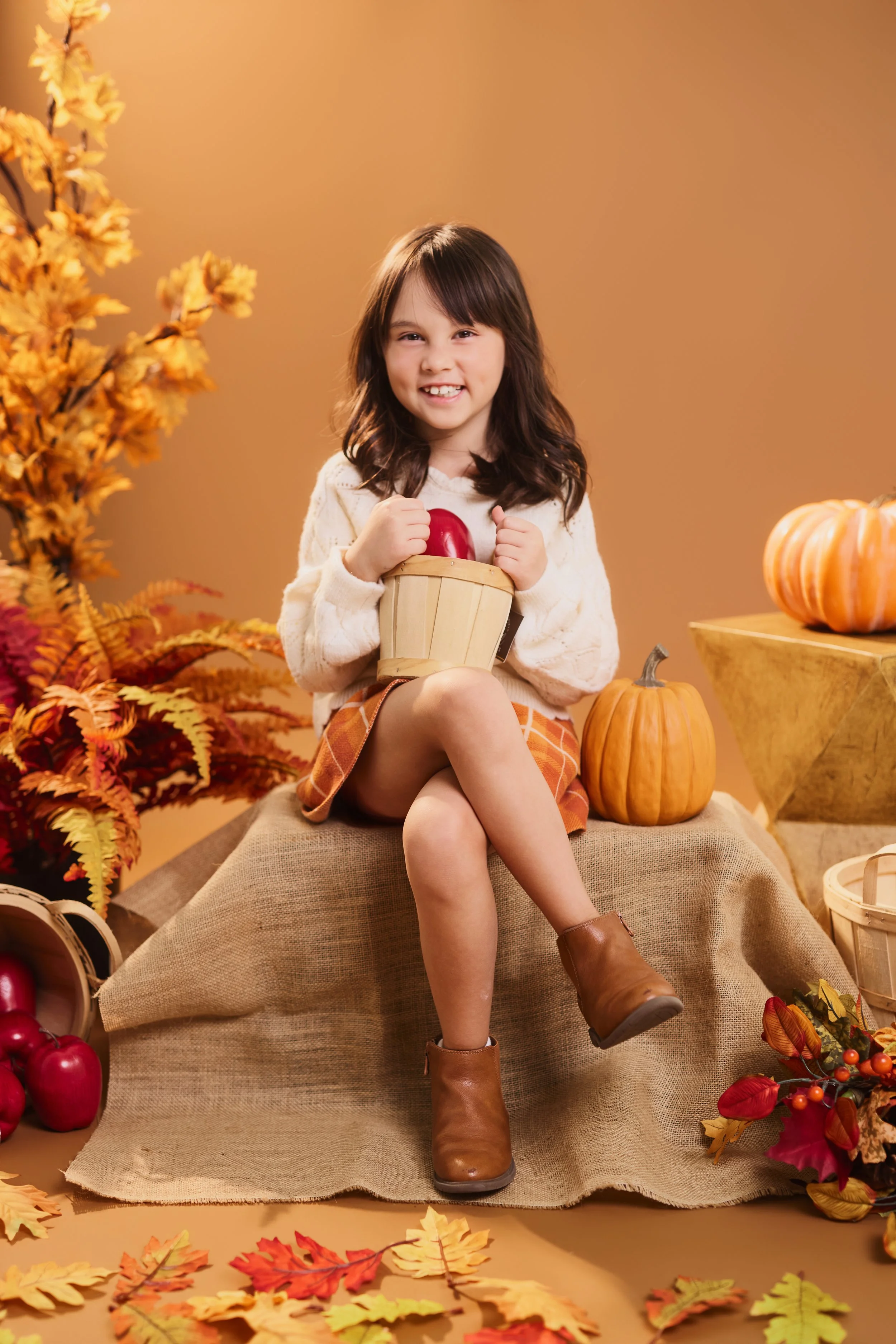 Young girl sitting on a burlap-covered platform surrounded by autumn leaves and pumpkins, holding a red bell pepper, smiling, in a fall-themed setting.