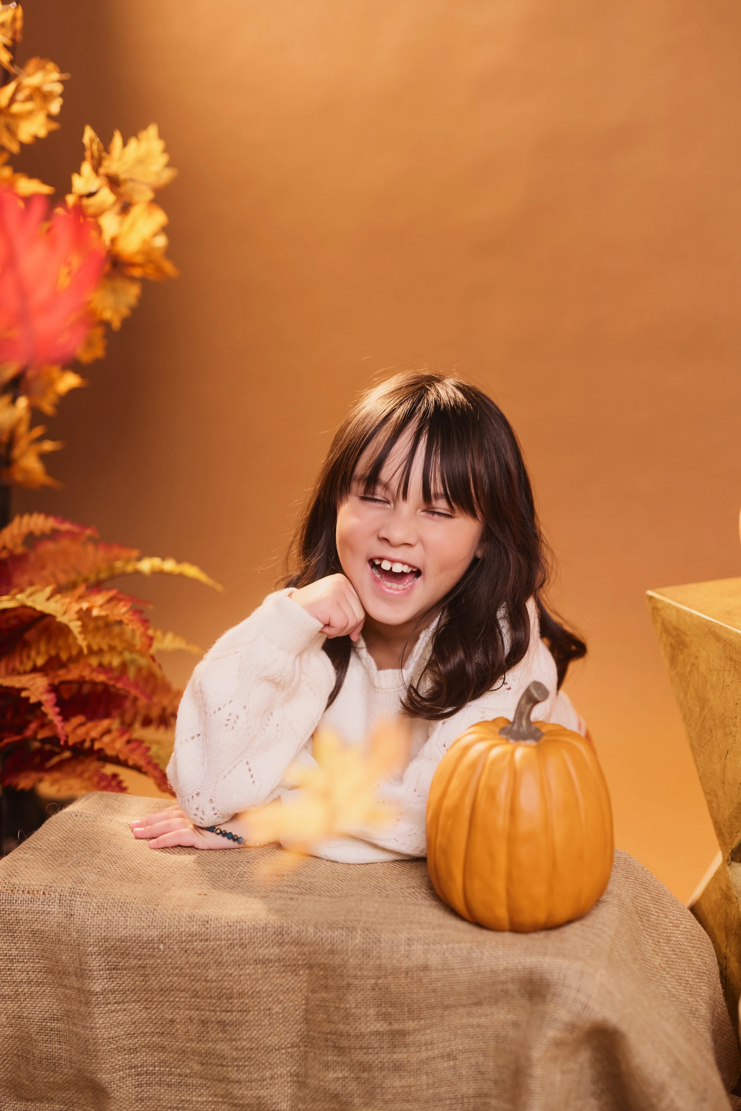 Smiling girl with dark hair and bangs, wearing a cream-colored sweater, sitting at a table with a pumpkin and fall decor.