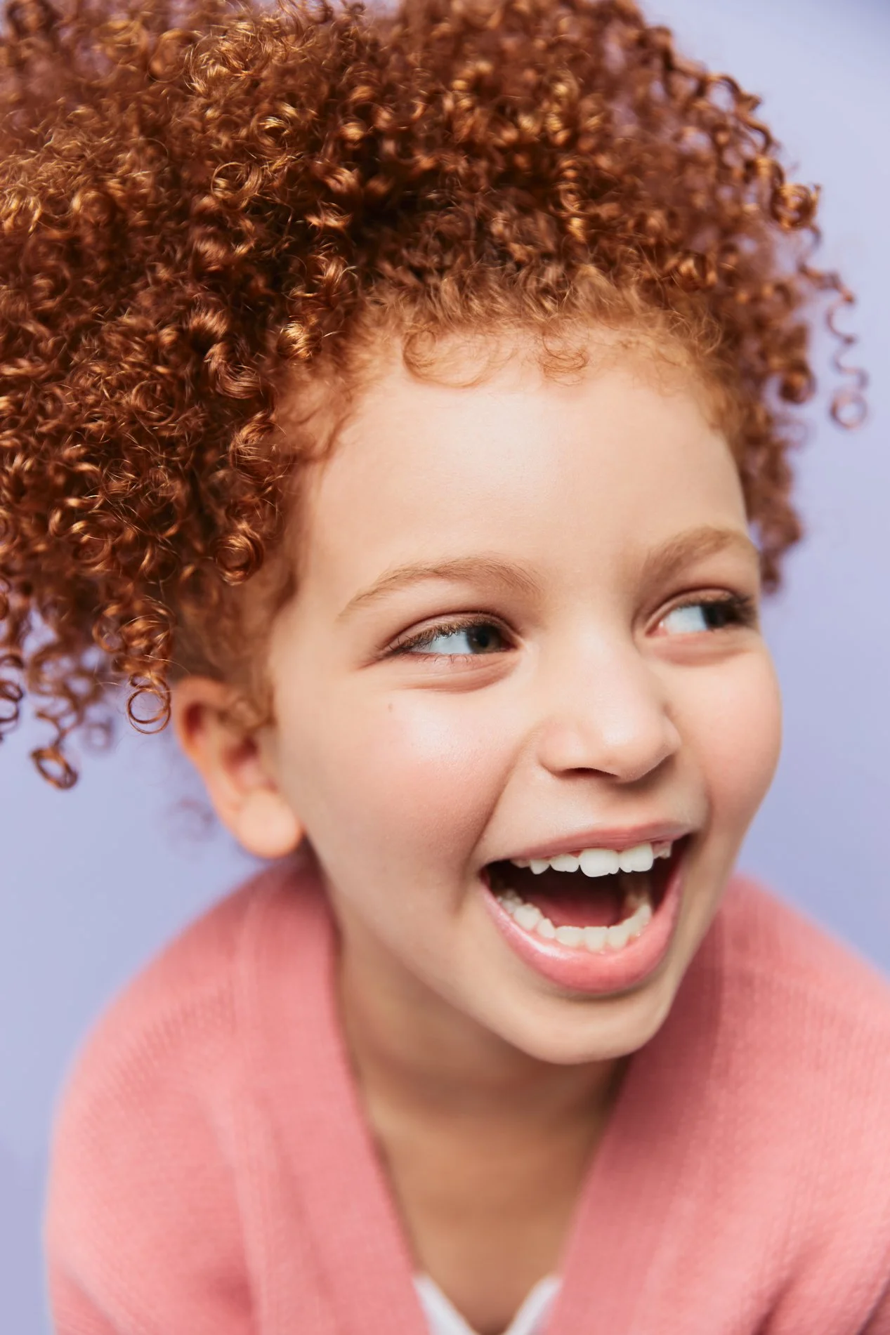 A young girl with curly red hair, smiling and laughing, wearing a pink sweater against a light purple background.