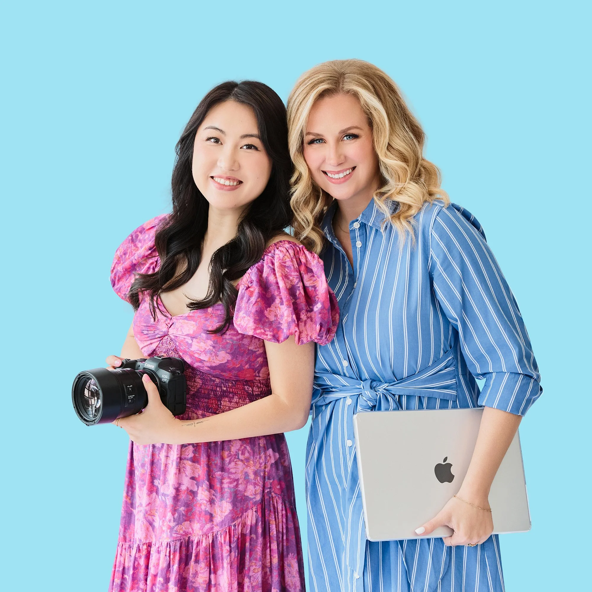 Two women standing close together smiling at the camera against a light blue background. One woman has long dark hair, wearing a pink patterned dress with holding a camera. The other woman has long blonde curly hair, wearing a blue striped shirt, holding a silver MacBook.
