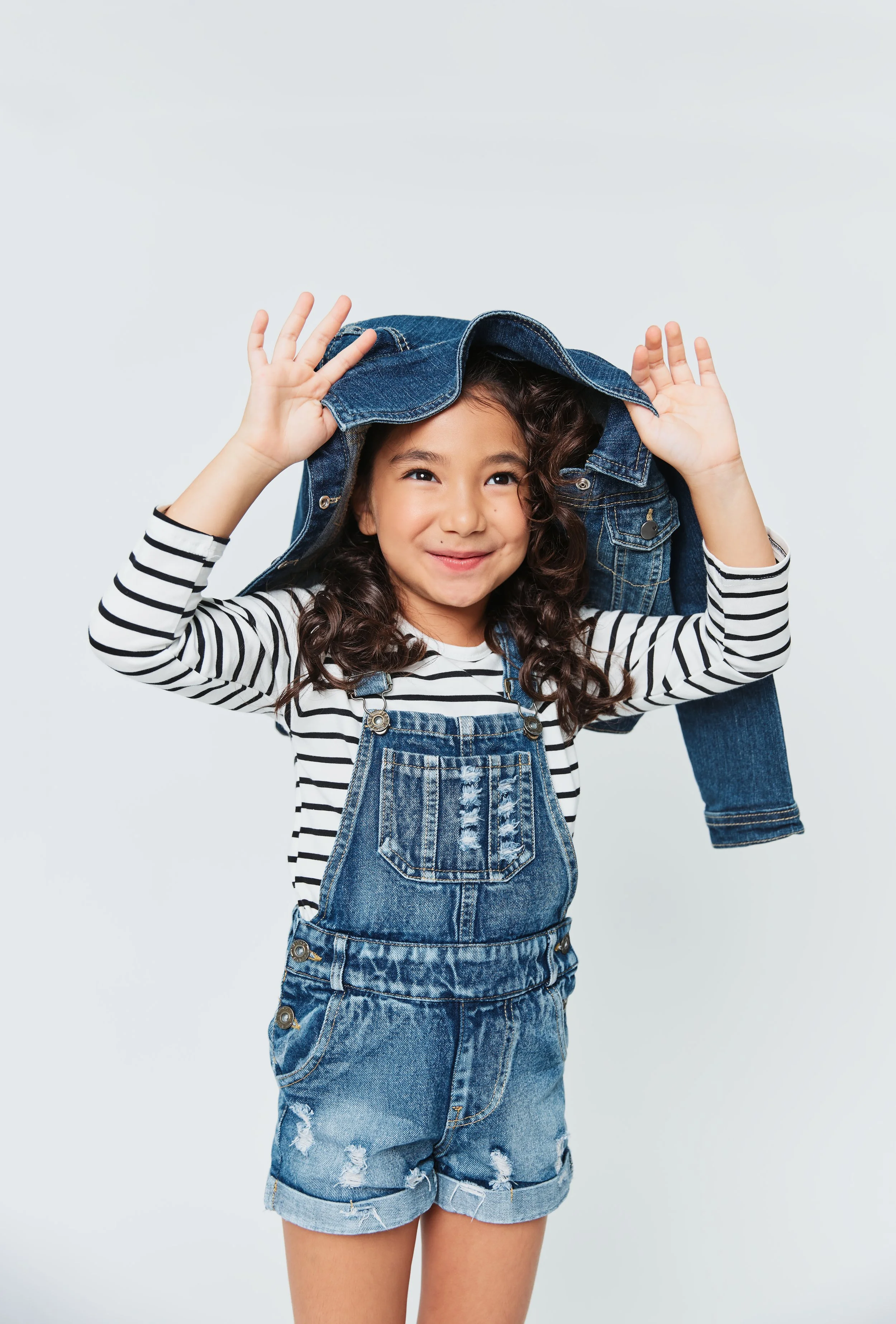 Young girl with curly hair smiling, wearing a black and white striped long sleeve shirt, denim overalls, and holding a denim jacket above her head.