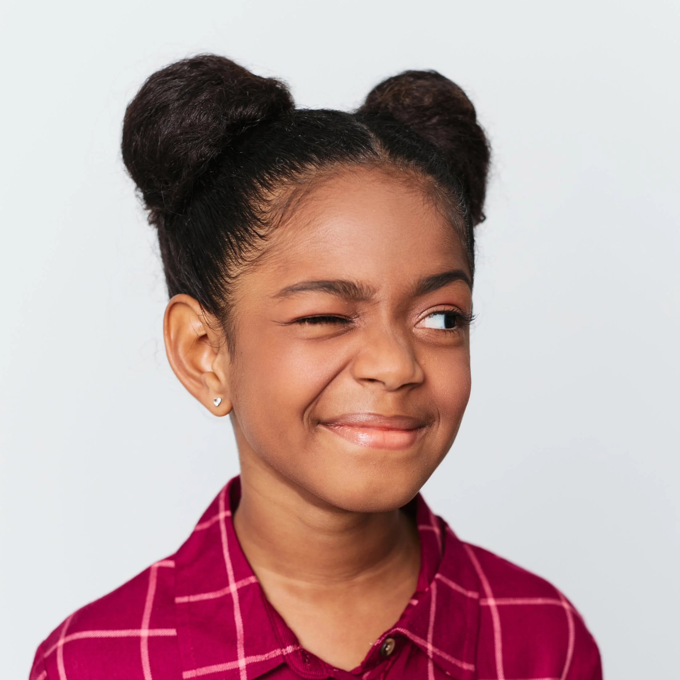 Close-up of a woman with dark skin and dark hair styled in two puffs, wearing a pink plaid shirt, winking and smiling.