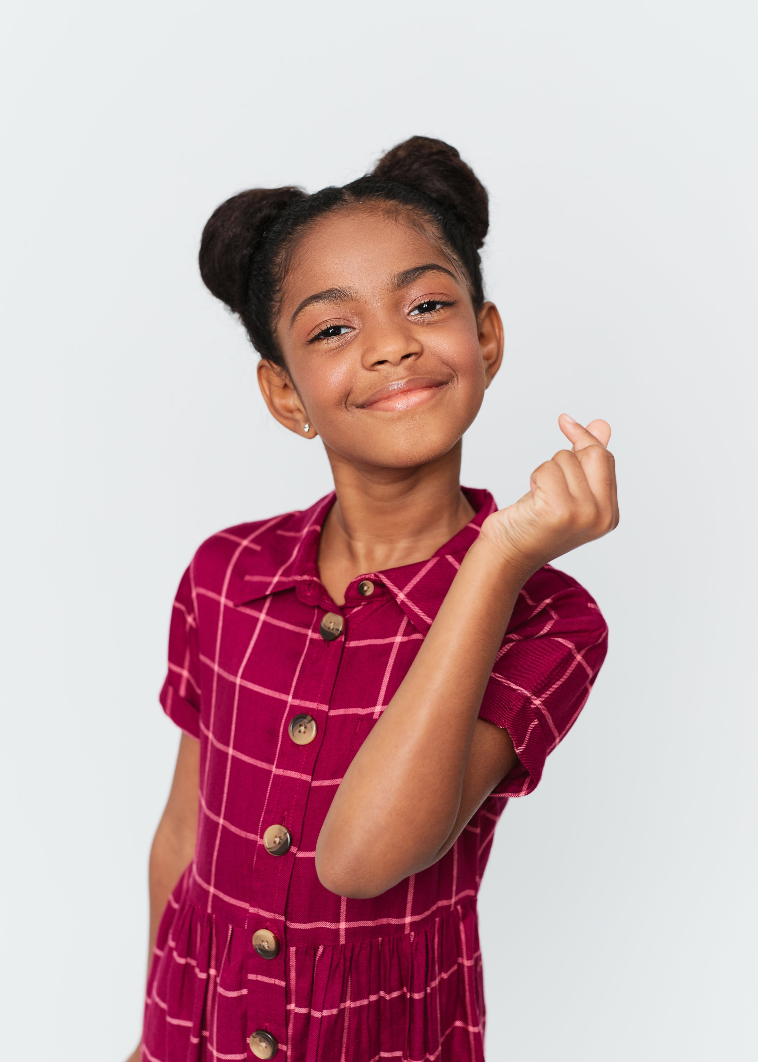 Smiling young girl with braided hair styled in two buns, wearing a maroon checkered dress, making a heart gesture with her hand, against a plain white background.