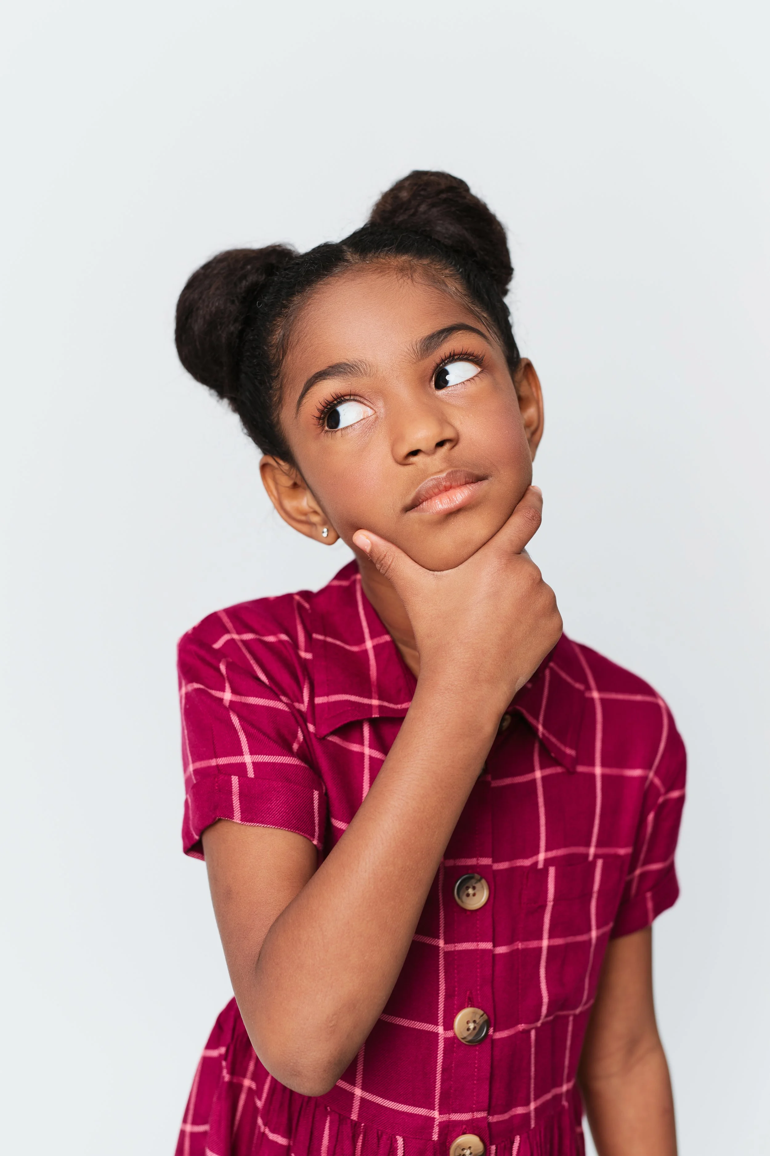 A young girl with dark hair styled in two buns, wearing a red and pink plaid dress, looks to the side with her hand on her chin, in front of a plain light background.