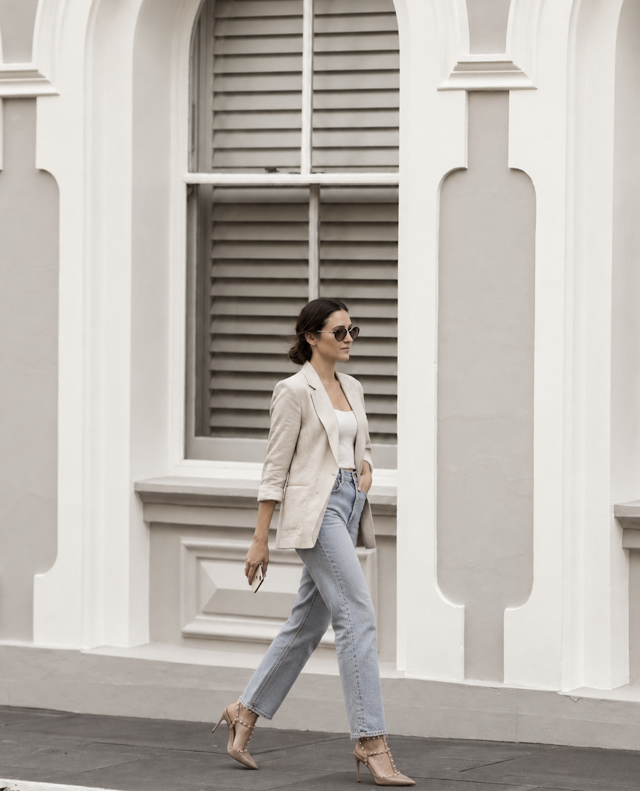 A woman with short dark hair wearing sunglasses, a beige blazer, white top, light blue jeans, and nude high heels, walking past a white building with decorative architectural details and large window shutters.
