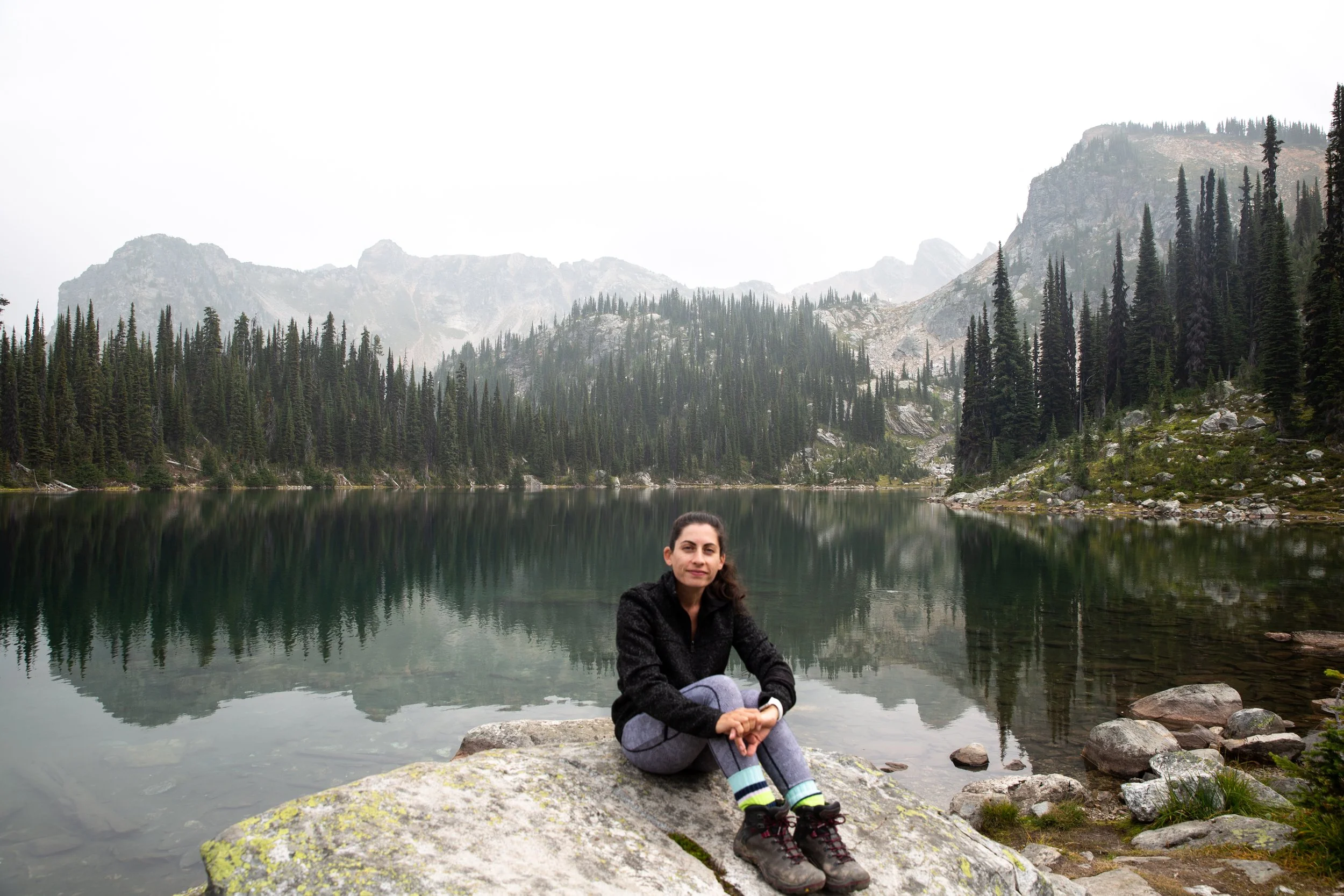A woman sitting on rocks by a calm lake with mountains and evergreen trees in the background.