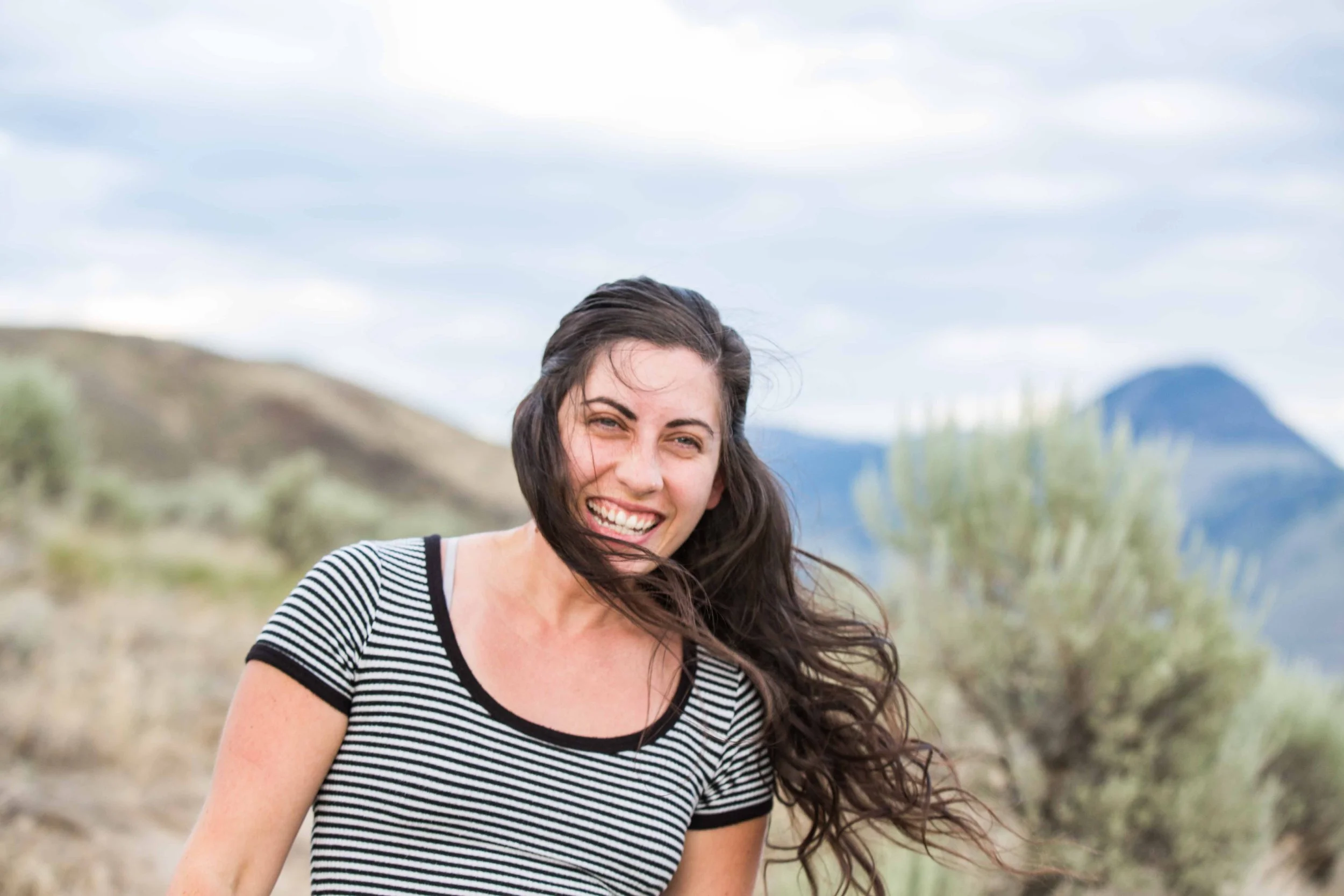 A woman with long dark hair smiling outdoors in a landscape with mountains and cloudy sky.