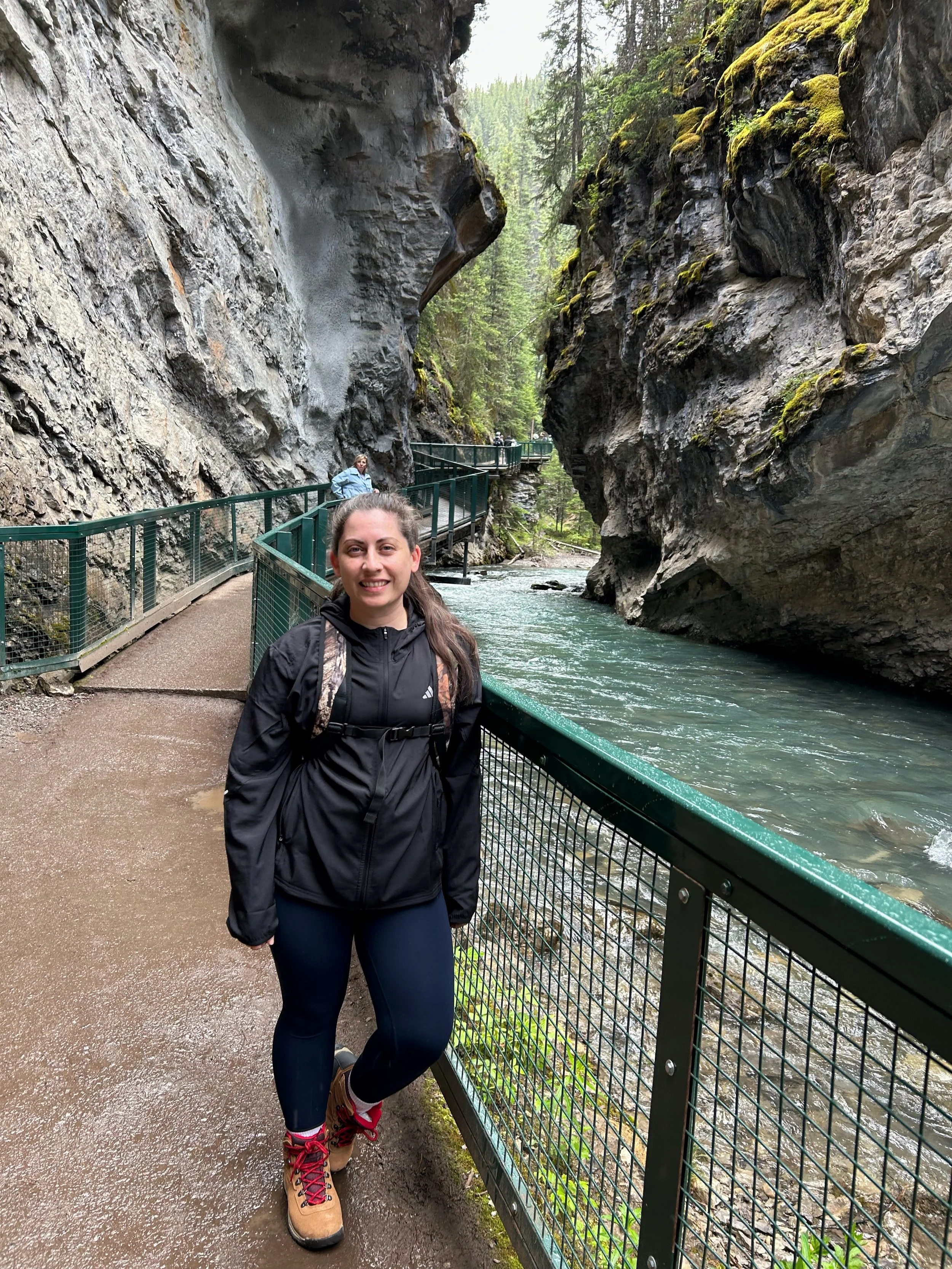 A woman with a backpack and hiking boots standing on a pathway next to a river, with a rocky canyon and dense evergreen forest in the background.