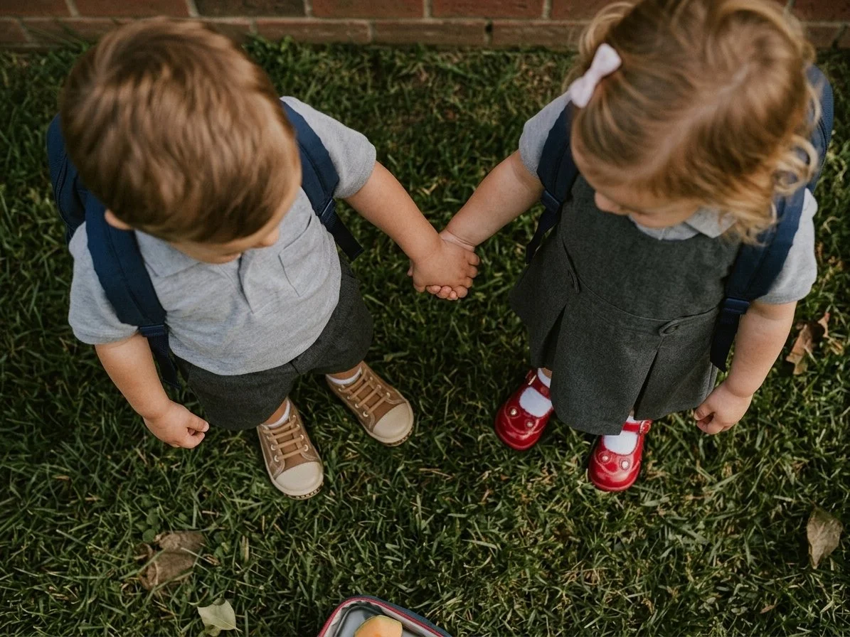 Overhead view of two young children wearing backpacks holding hands on grass beside a brick wall