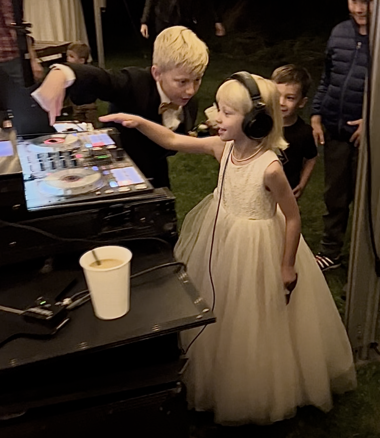 A young girl in a fancy, light yellow dress with a full skirt and sleeveless top, wearing large headphones, is dancing and smiling in front of a DJ setup at a party. Several people are standing and watching her.