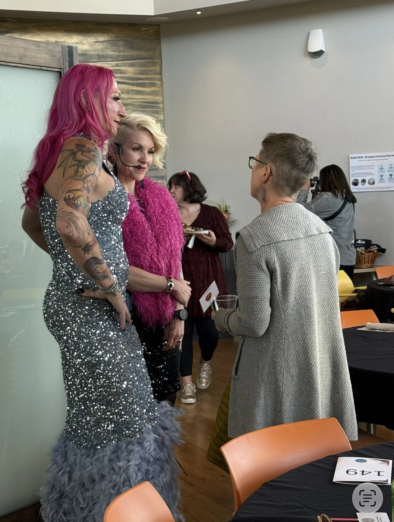 Two women with colorful hair and glamorous outfits talking to a young woman with glasses, in a room with tables and chairs, at an event or gathering.