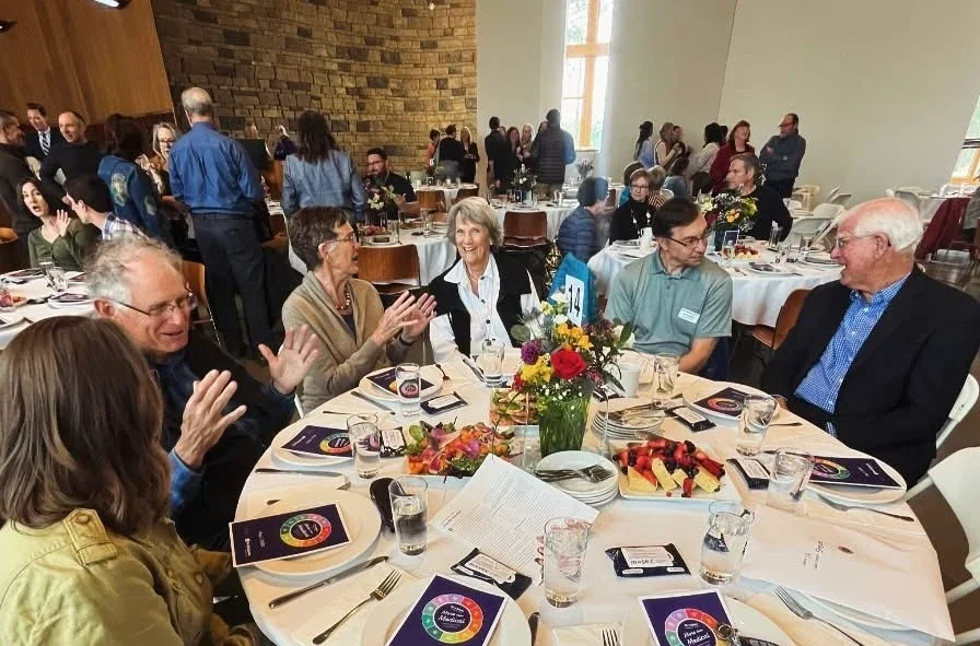 People sitting at round tables, engaging in conversation, at a formal event or banquet in a large room with high windows and wood-paneled wall.