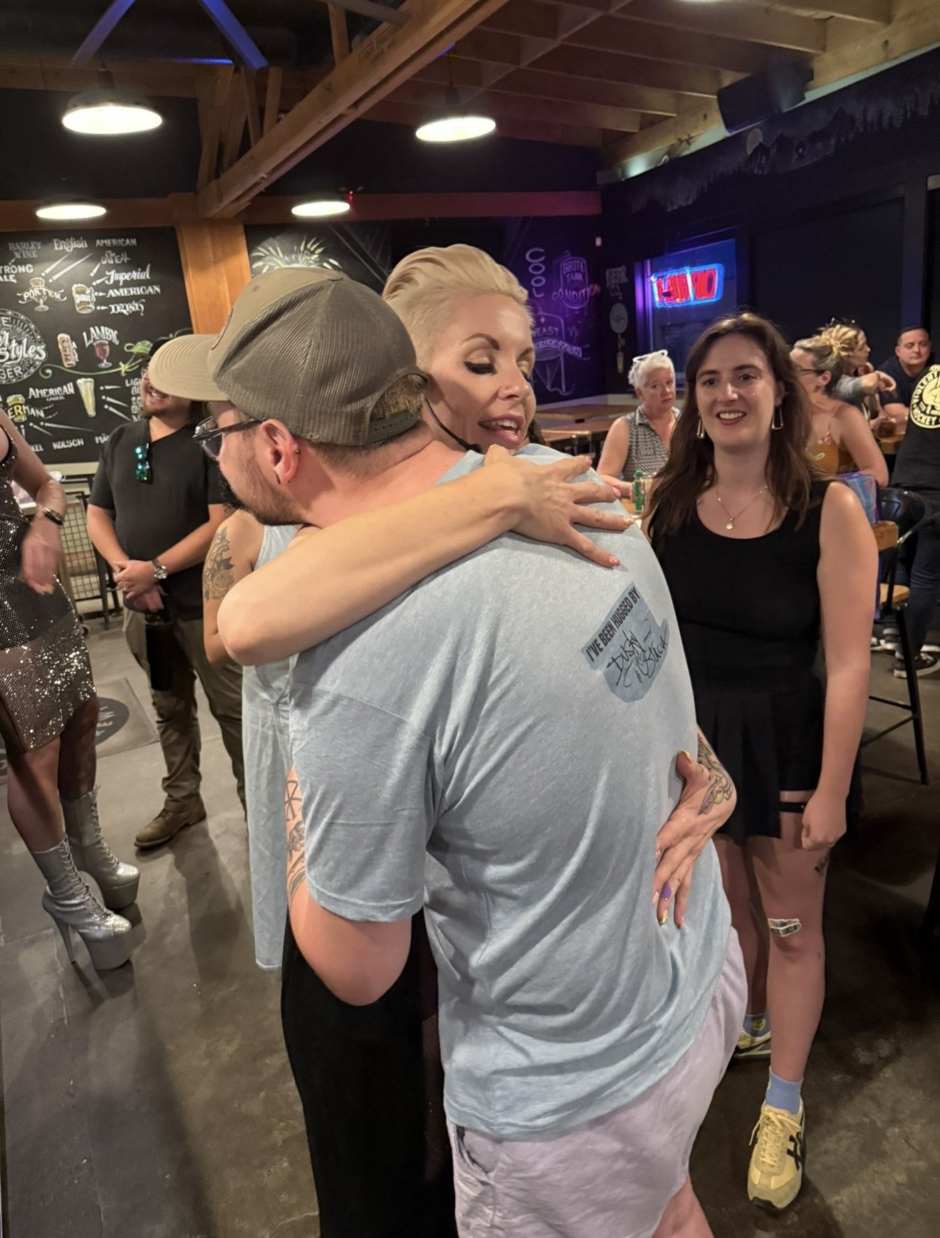People hugging and socializing in a lively bar or restaurant with blackboard menu and neon signs in the background, and a diverse group of patrons.