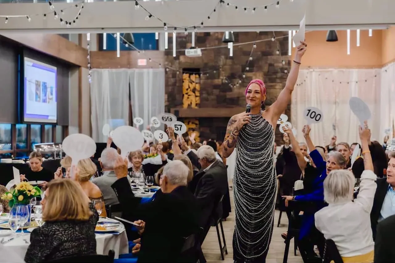 A woman with pink hair holding a microphone and raising her hand at a crowded dinner event with auction paddle cards.
