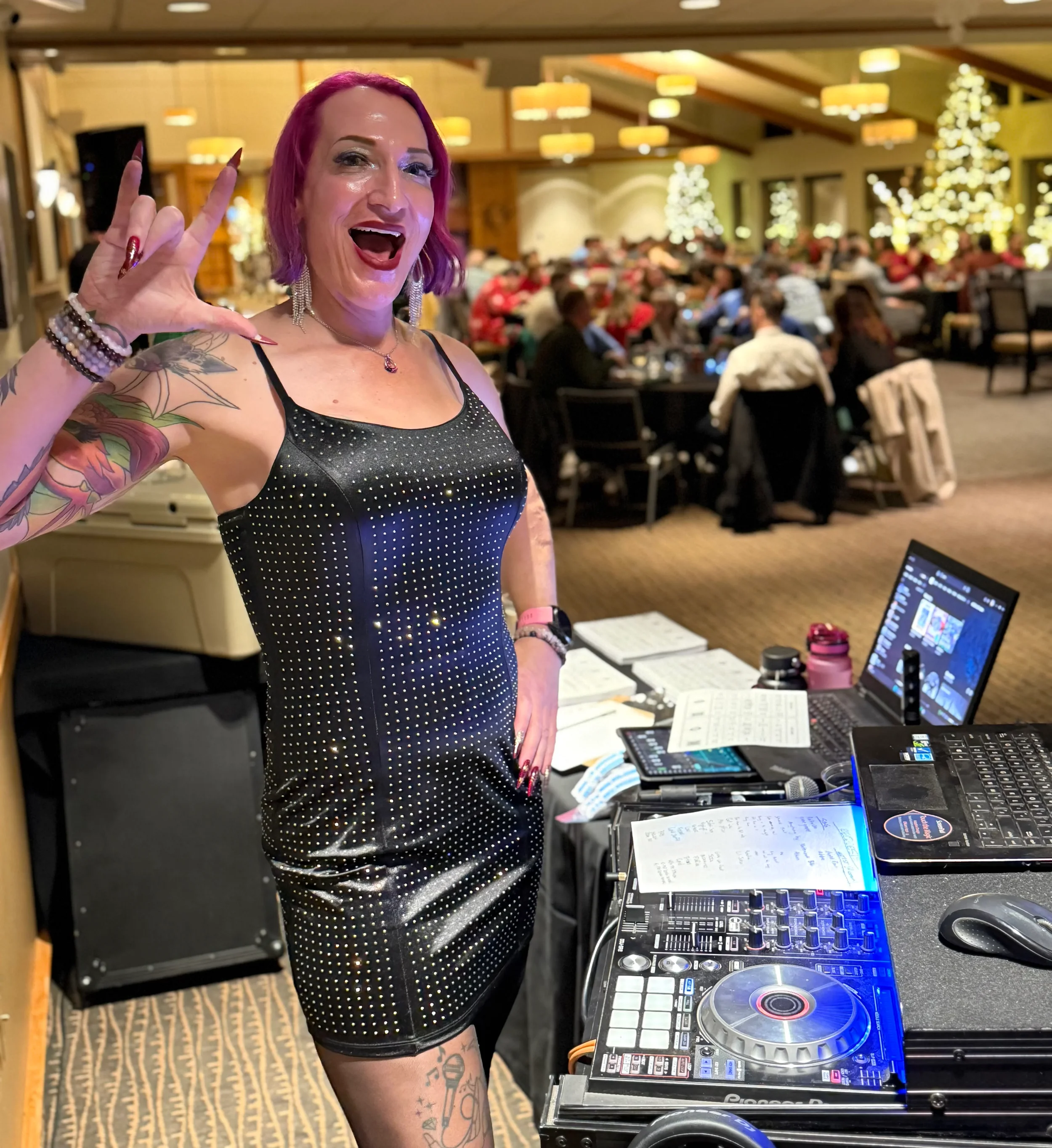 A woman with pink hair and tattoos, smiling and making the 'I love you' hand gesture at a DJ booth during a festive indoor event, with many people seated at tables and decorative Christmas trees in the background.