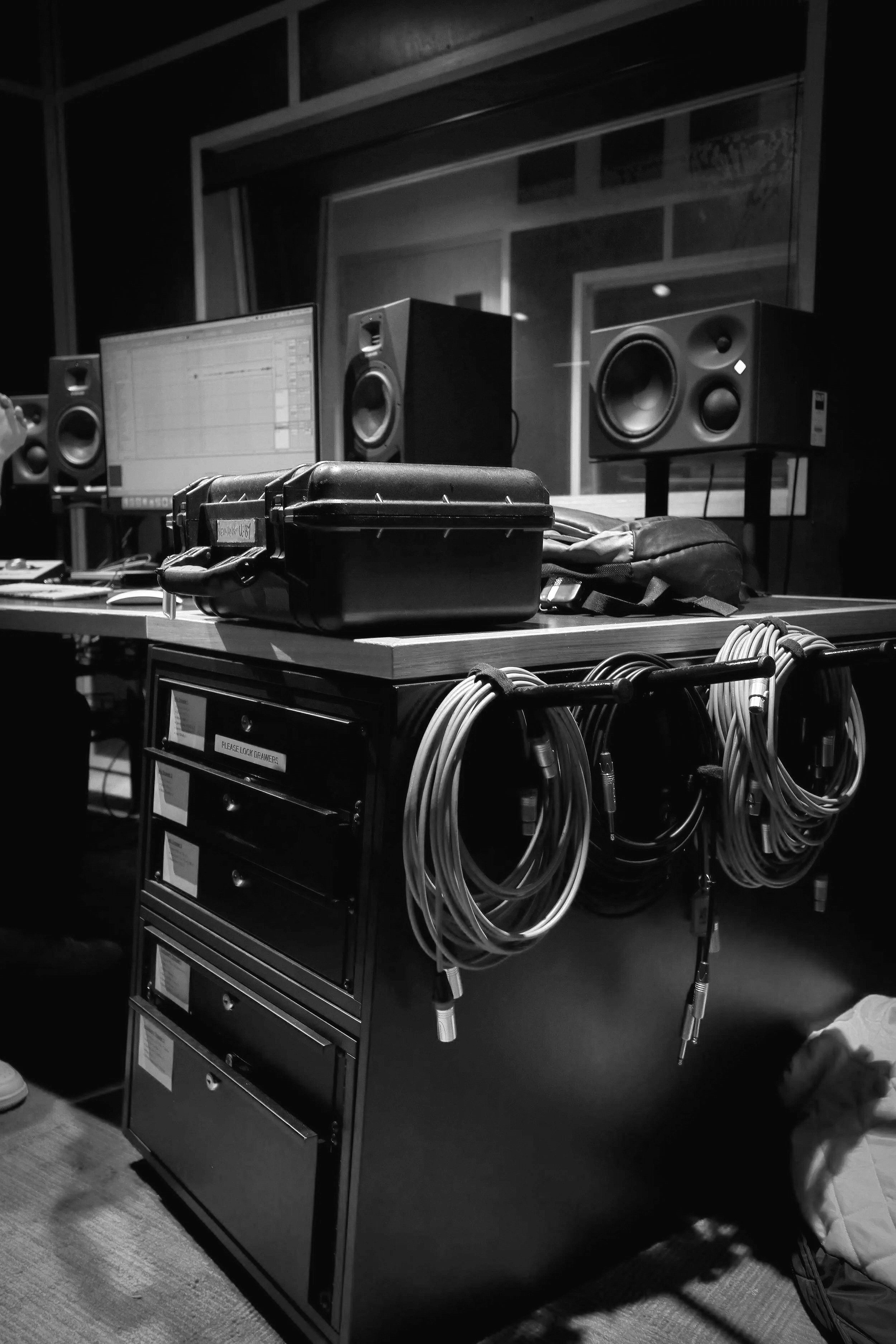 In a recording studio, there is a black cabinet with multiple drawers and a sign that says 'Please Lock Doors.' Several audio cables are hanging from the cabinet. In the background, there are two large studio monitors on stands, a computer monitor displaying a digital audio workstation, and soundproof walls.