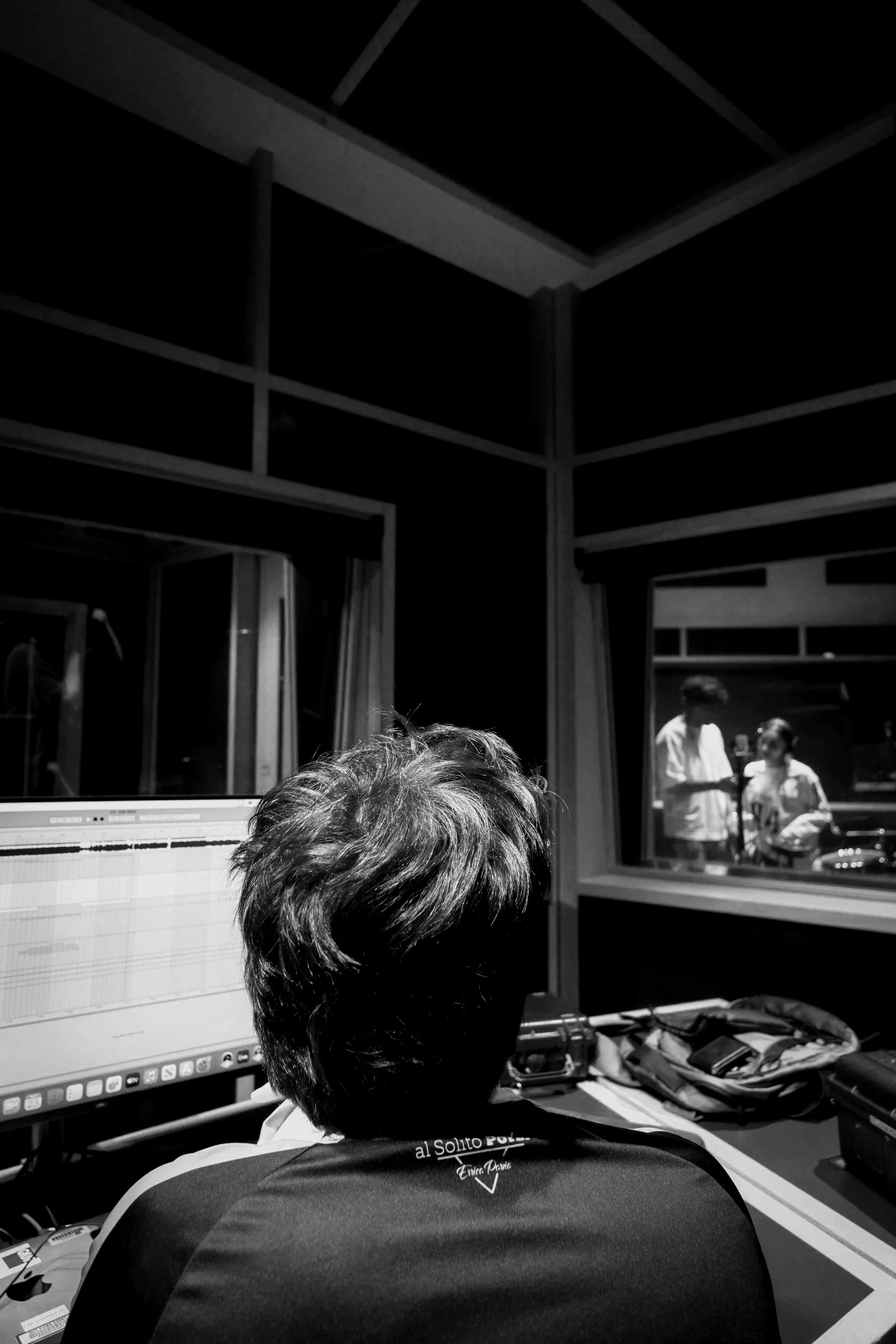 A person with dark hair is sitting in front of a computer monitor displaying an audio editing software. In the background, through a window, two local Brisbane based artists are engaged in discussion.