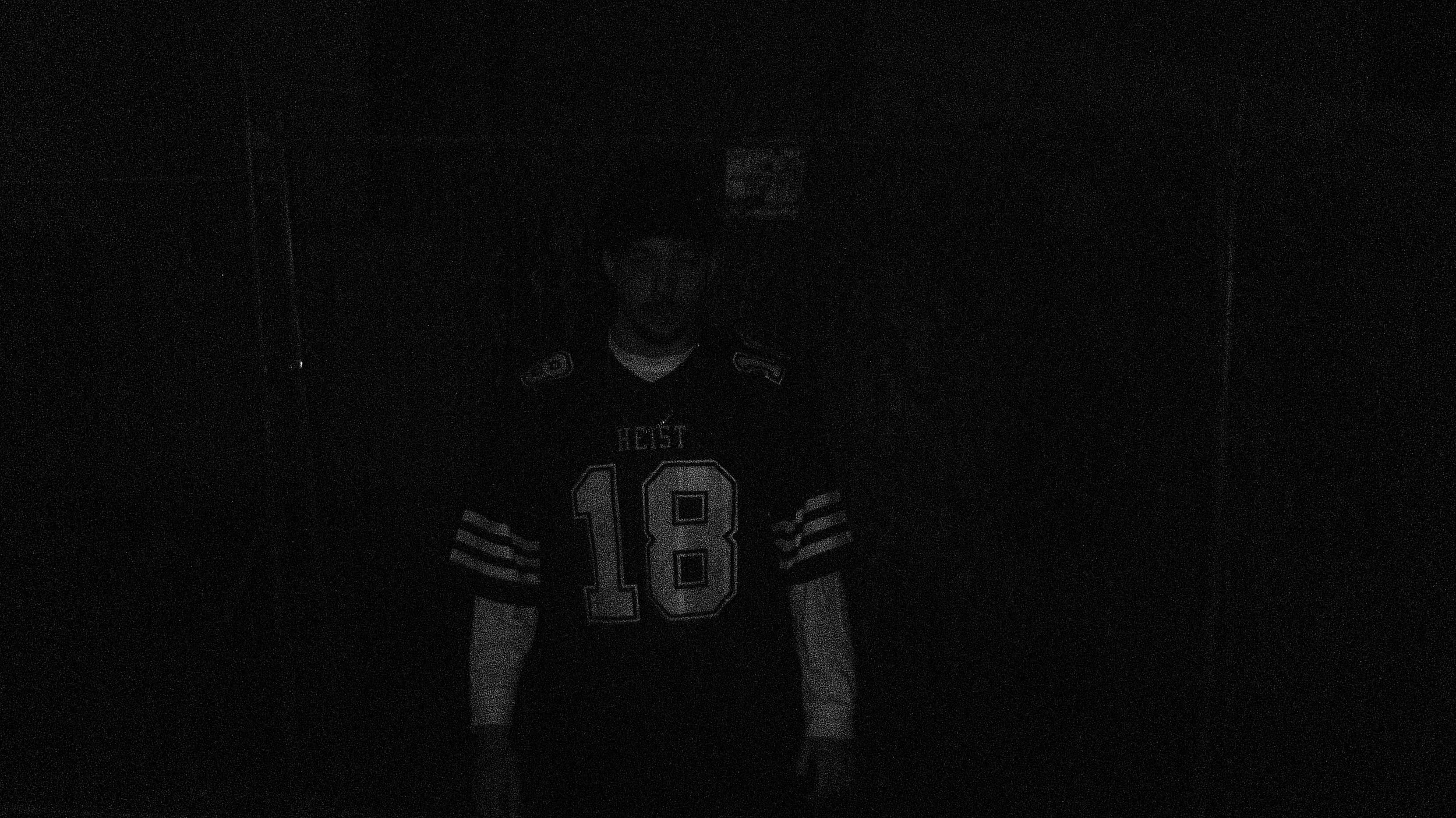 A man wearing a black sports jersey with the number 18 and the word 'HEIST' on it, standing in a dark room.