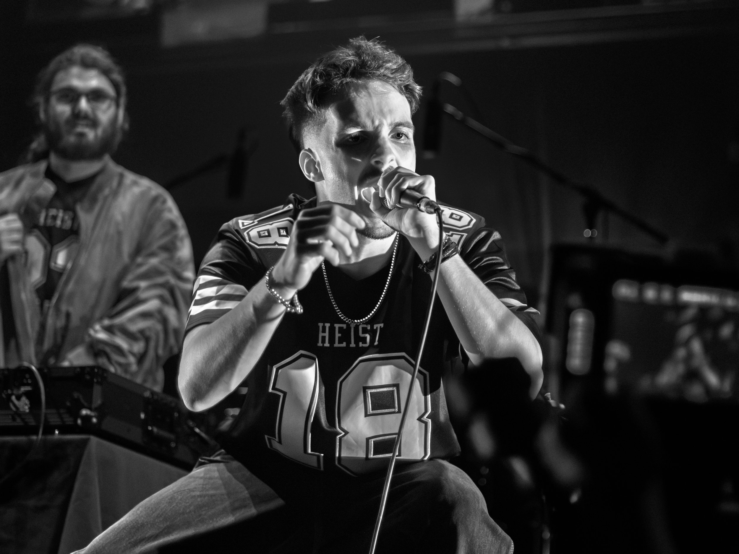 A black and white photo of a young man with styled hair, wearing a sports jersey with the number 18, rapping into a microphone during a performance or event at Gardens Theatre, Brisbane at the QUT Showcase, DJ Lyre in the background.