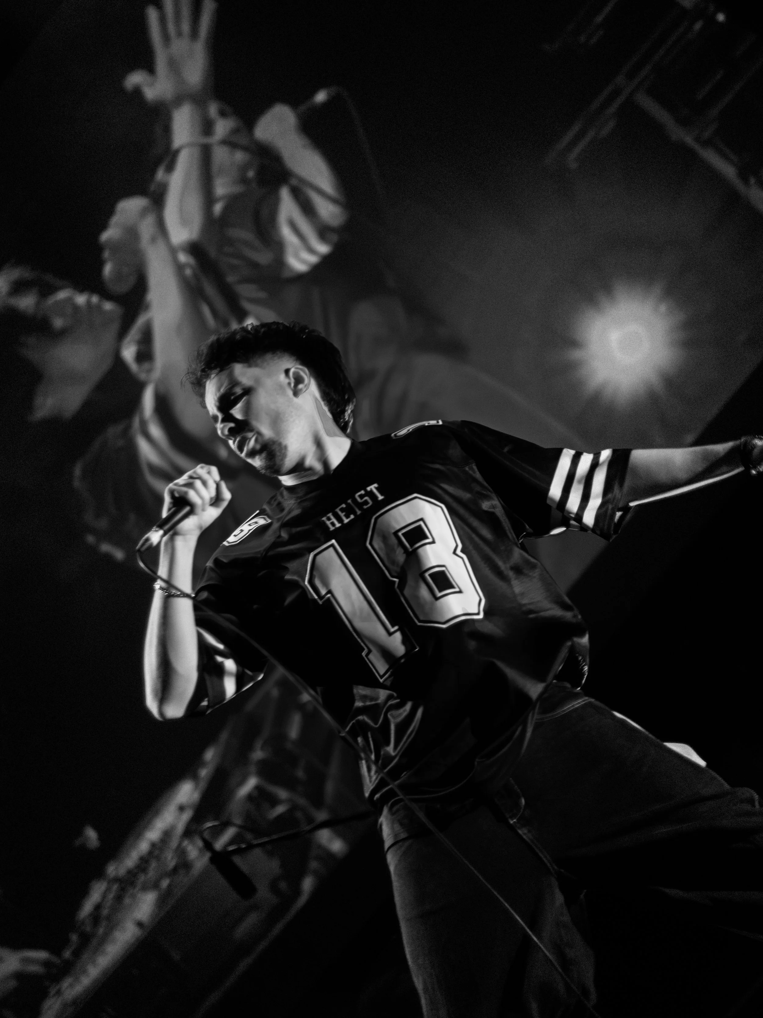 Local Brisbane Rapper performing at the QUT Showcase, holding a microphone in his right hand, wearing a sports jersey with the number 18 and the word 'HEIST' visible, with a large projection of a face in the background.