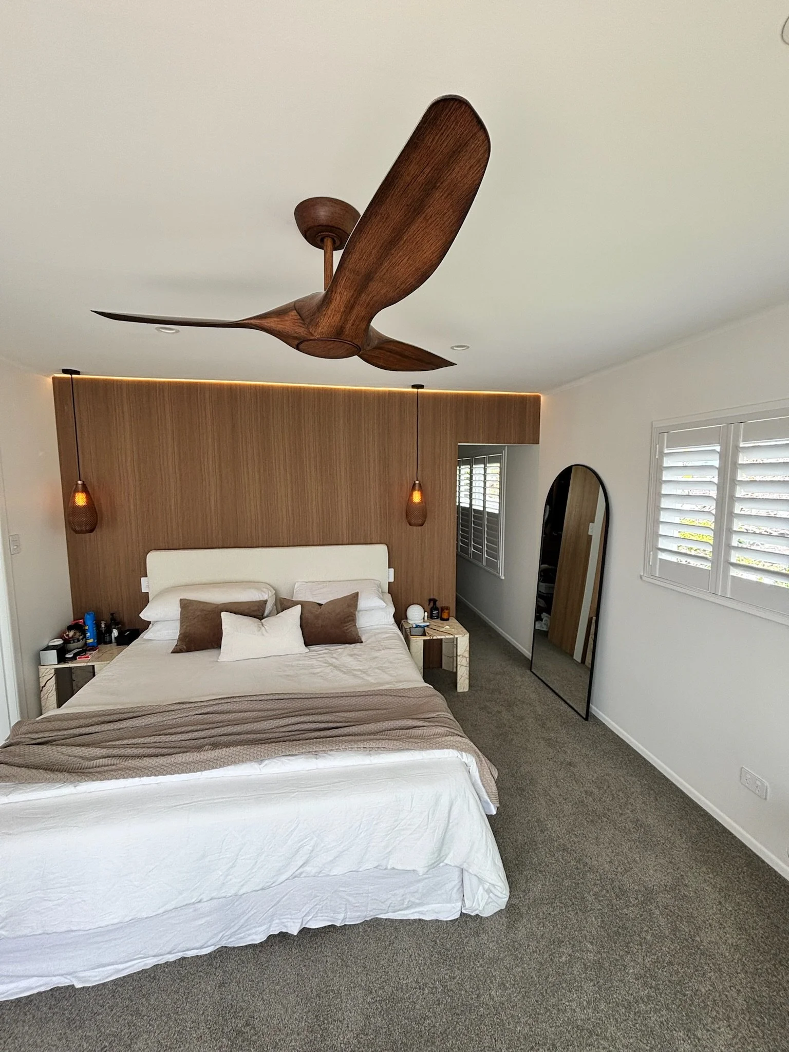 A bedroom with a bed, nightstands, a large mirror, and a ceiling fan. The bed has white sheets and pillows, and there are two hanging pendant lights on either side of the bed. The room has a window with white shutters.