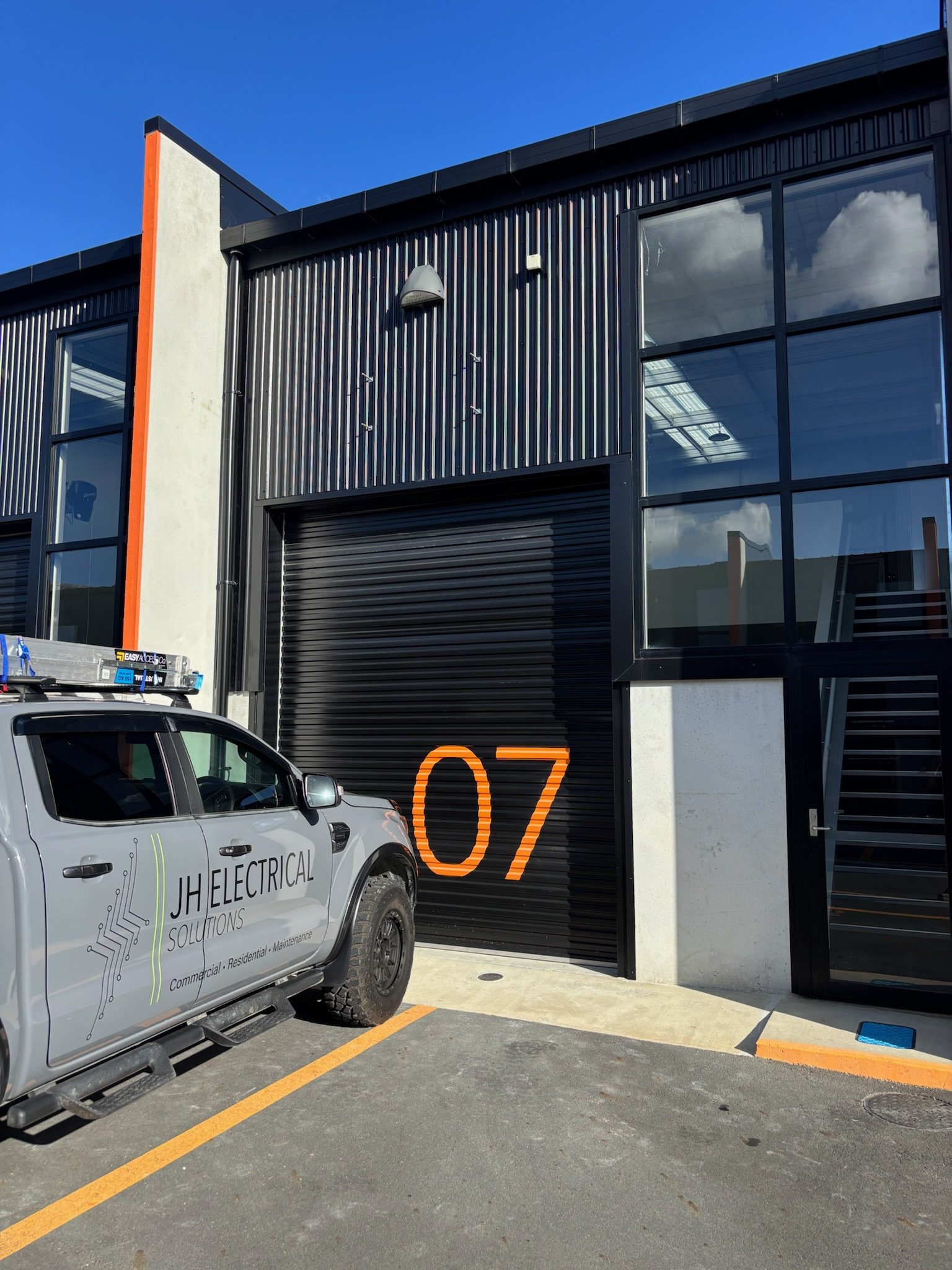 Modern commercial building with black metal siding, large glass windows, and an orange accent strip. A white utility vehicle with the logo 'JH Electrical Solutions' is parked in front. The sky is clear and blue.
