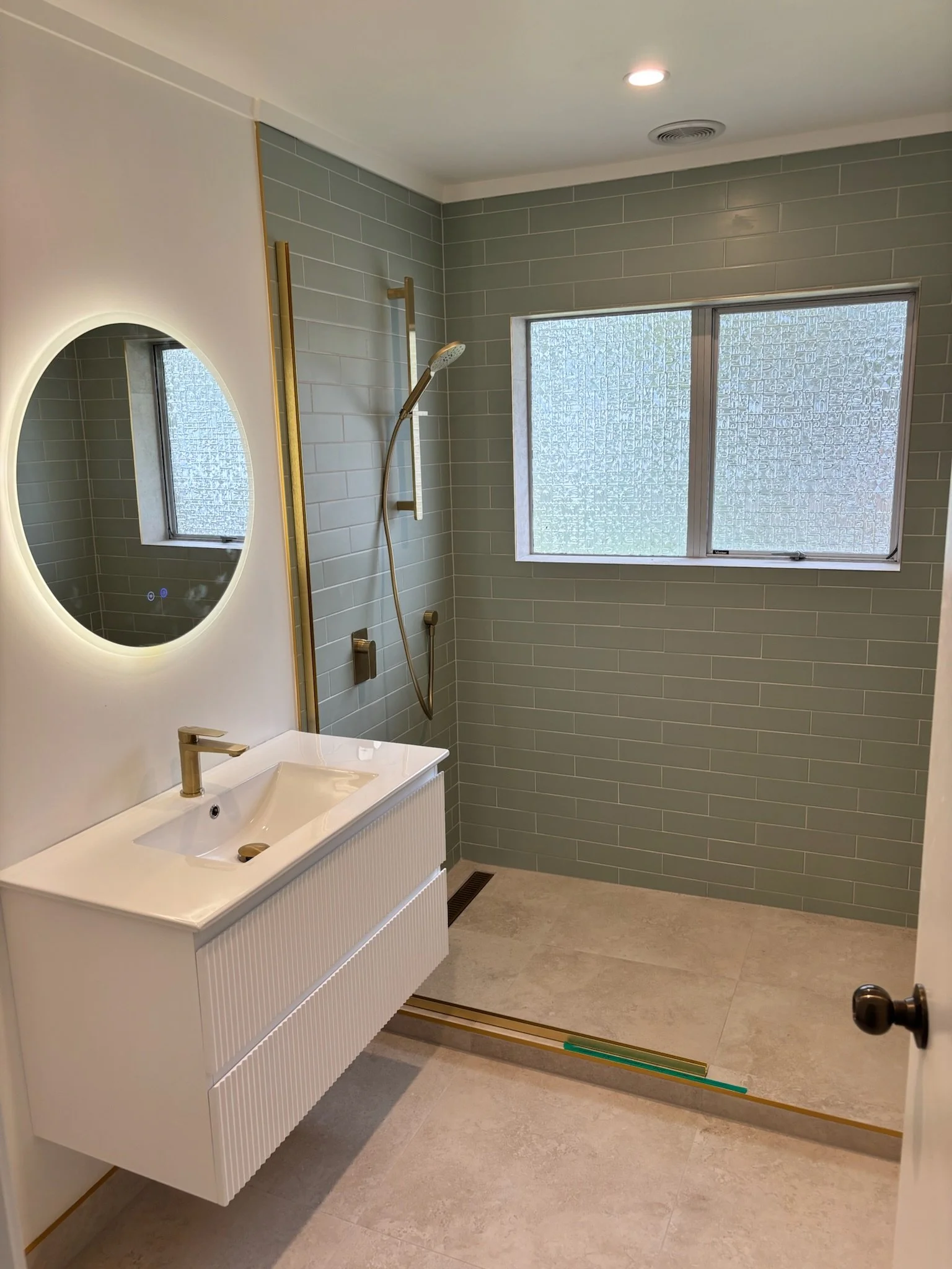 Modern bathroom with green tiled wall, frosted window, white vanity with a sink, round mirror with backlight, and shower area with gold fixtures and frosted window.