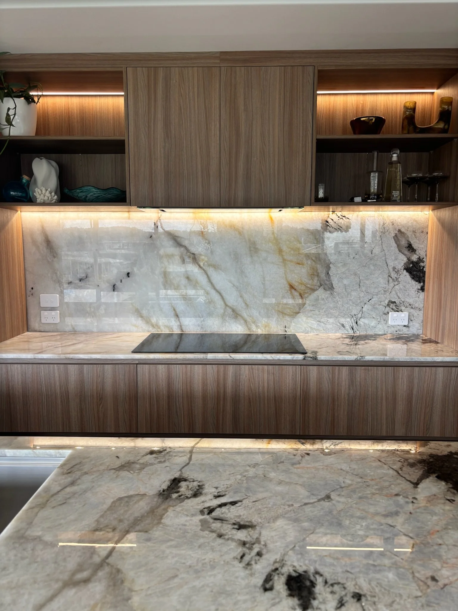 A kitchen backsplash made of large marble slabs with gray, black, and gold veining, above a matching marble countertop with similar veining, and wooden cabinets with built-in LED lighting.