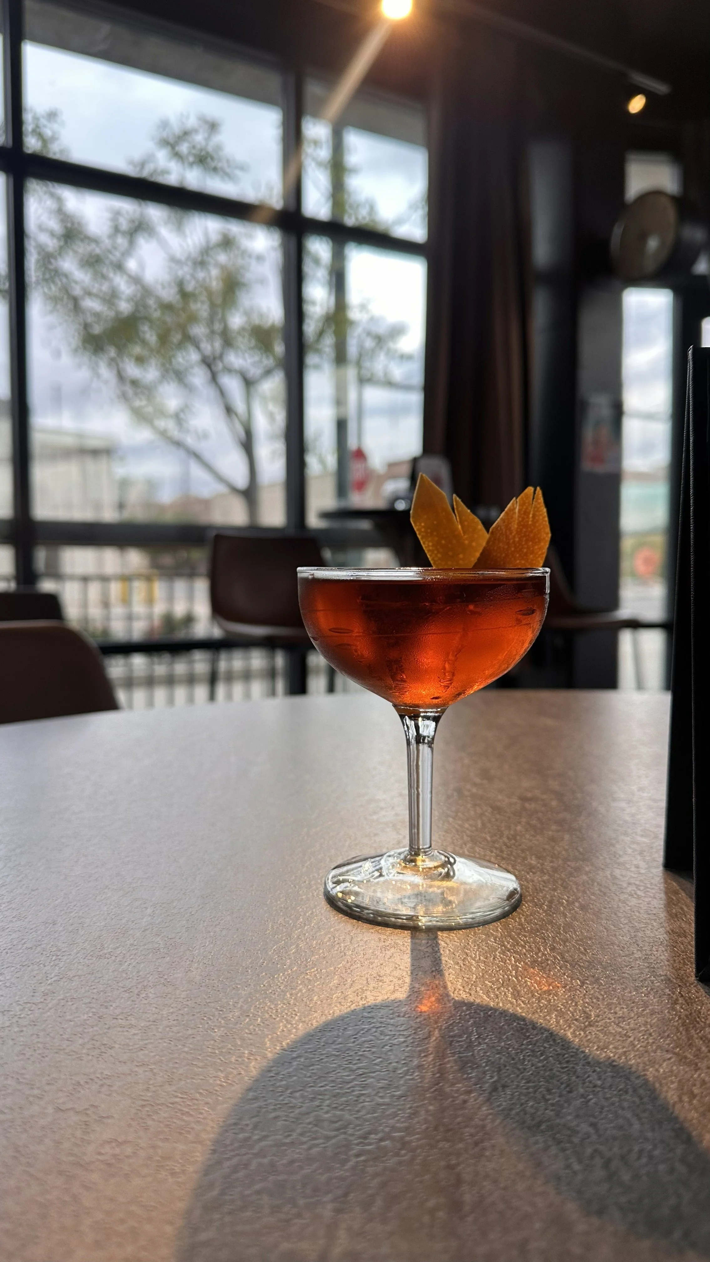 A cocktail glass with a pinkish-red drink garnished with orange peel, placed on a table inside a restaurant with large windows and a tree visible outside.