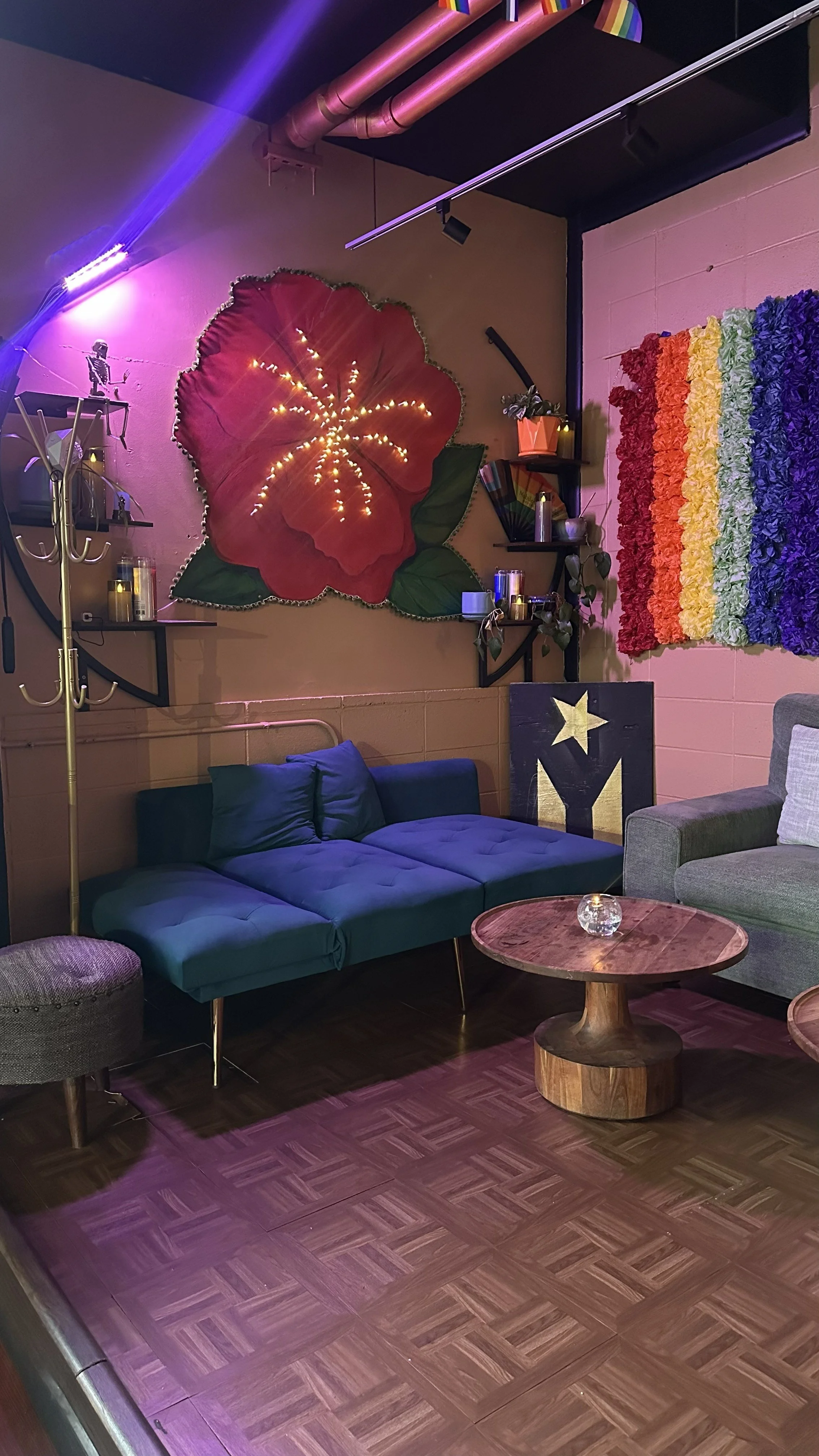 A cozy living room with a blue couch, a gray armchair, and a wooden coffee table. Decor includes a large red flower wall art with lights, a rainbow-colored wall hanging, a small shelf with plants and candles, and a black artwork with a star. The room has dark wooden flooring and a purple light source.