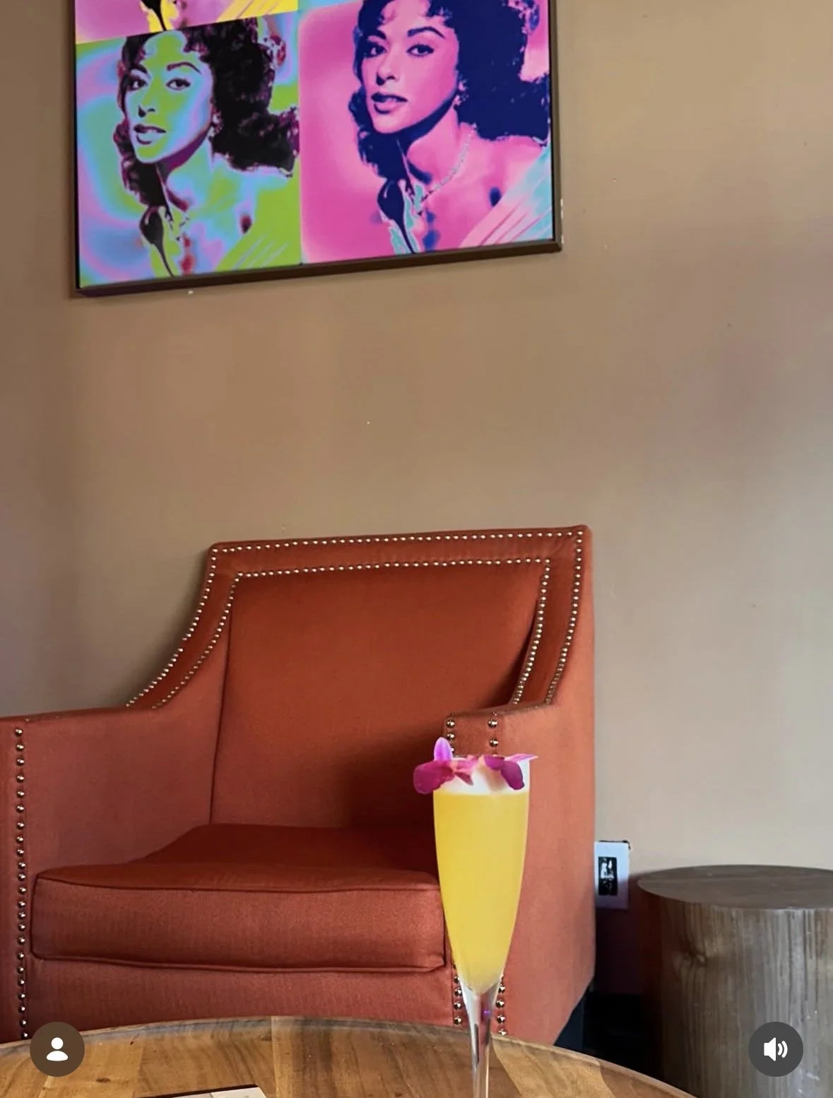 A tropical yellow cocktail with purple flower garnishes in a tall glass on a wooden table. Behind it is an orange armchair with nailhead trim, against a beige wall. A framed license plate art with portraits of women in vibrant colors hangs on the wall above.