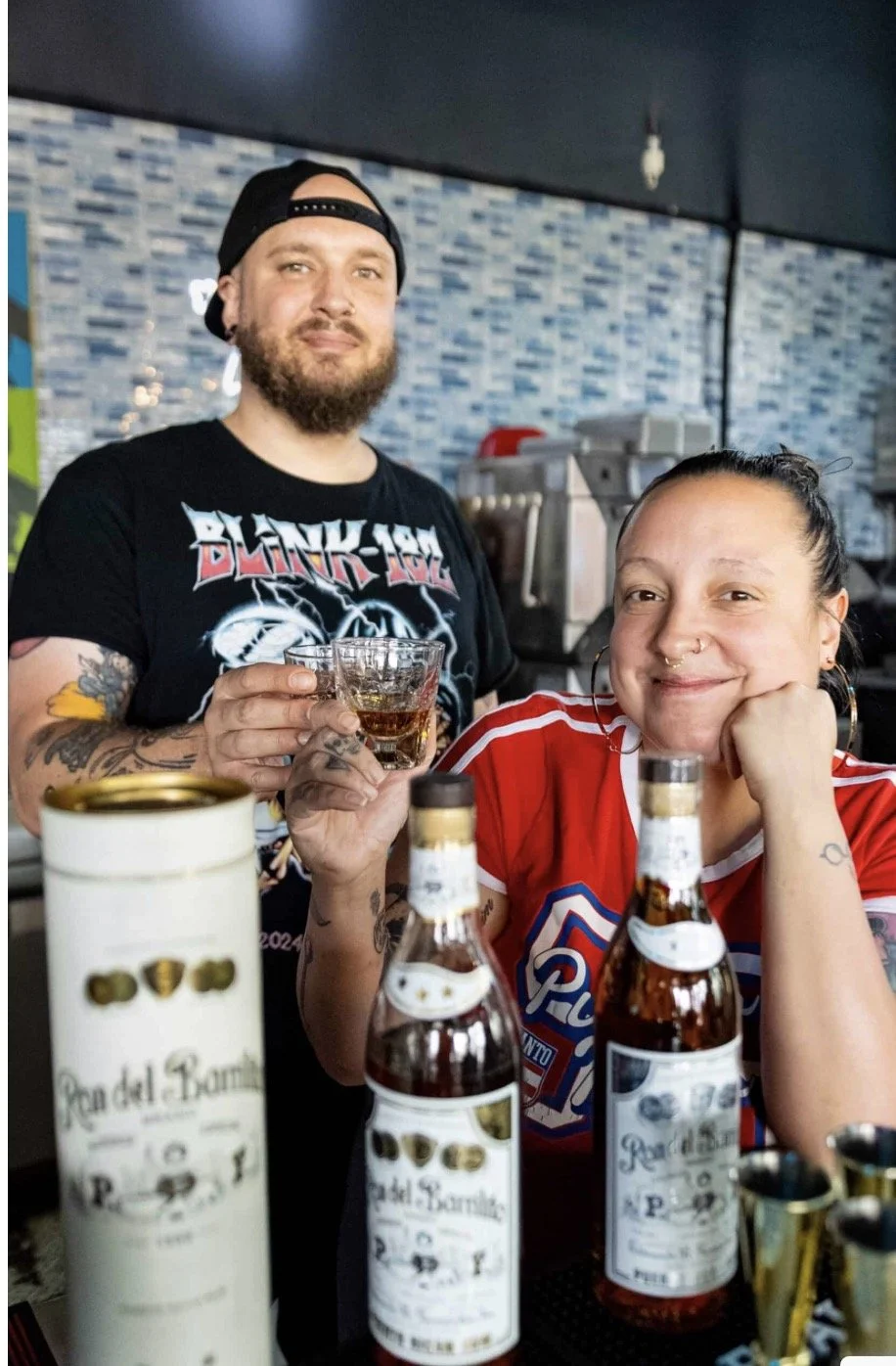 A man with a beard and tattoos, wearing a Black Label shirt and backward cap, holds a glass of liquor. A woman with dark hair tied back, wearing a red sports jersey, sits with her chin resting on her hand, smiling. Two bottles of alcohol and a cylindrical container are on the counter in front of them, with a blue mosaic wall in the background.