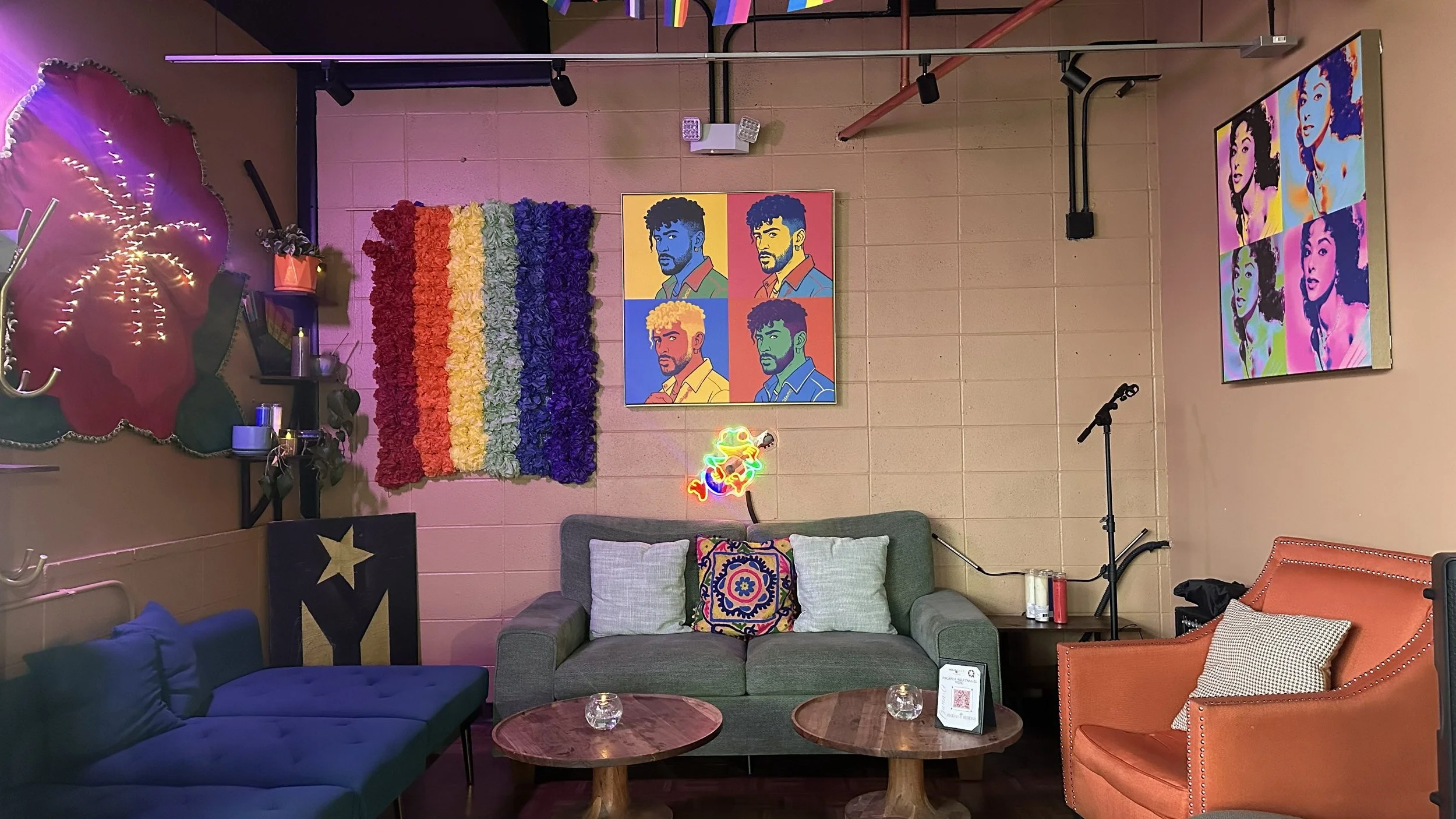 An interior scene of a cozy living room decorated with colorful artwork and furniture, including a gray sofa with patterned cushions, an orange armchair, and a coffee table. The walls feature pop art portraits and a rainbow-colored floral piece, with neon signs and string lights adding to the vibrant atmosphere.