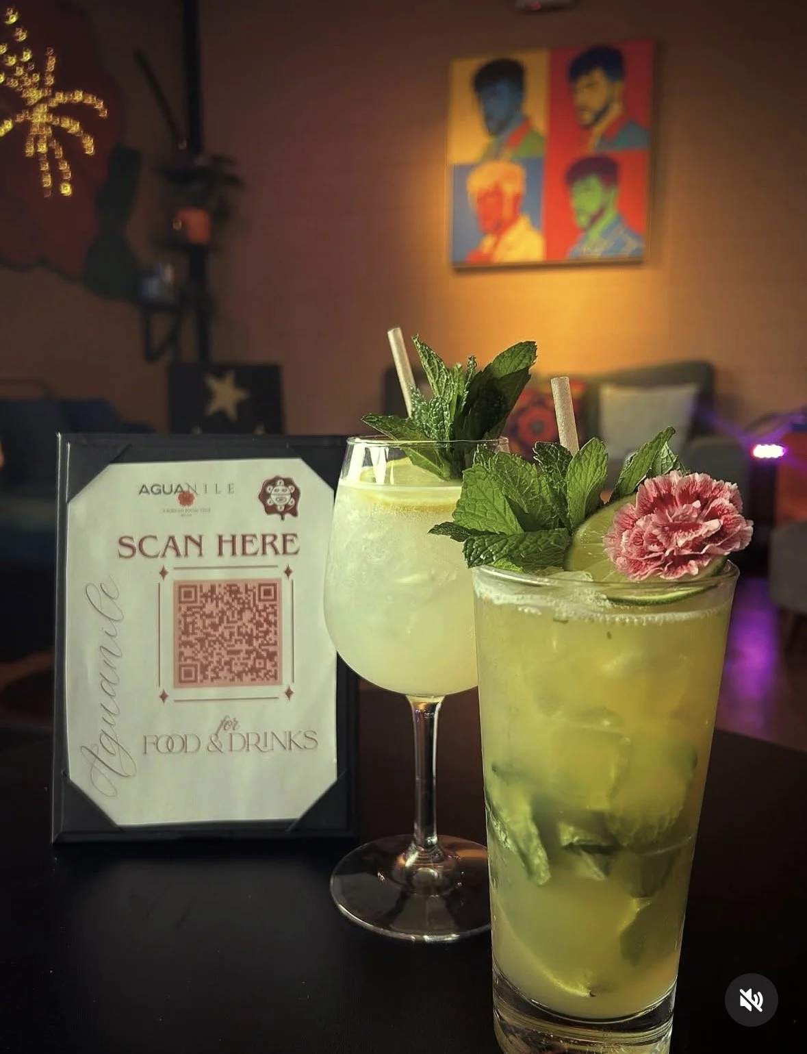 Two mojito cocktails garnished with mint, lime, and flower, with a QR code sign on a table in a dimly lit bar or restaurant.