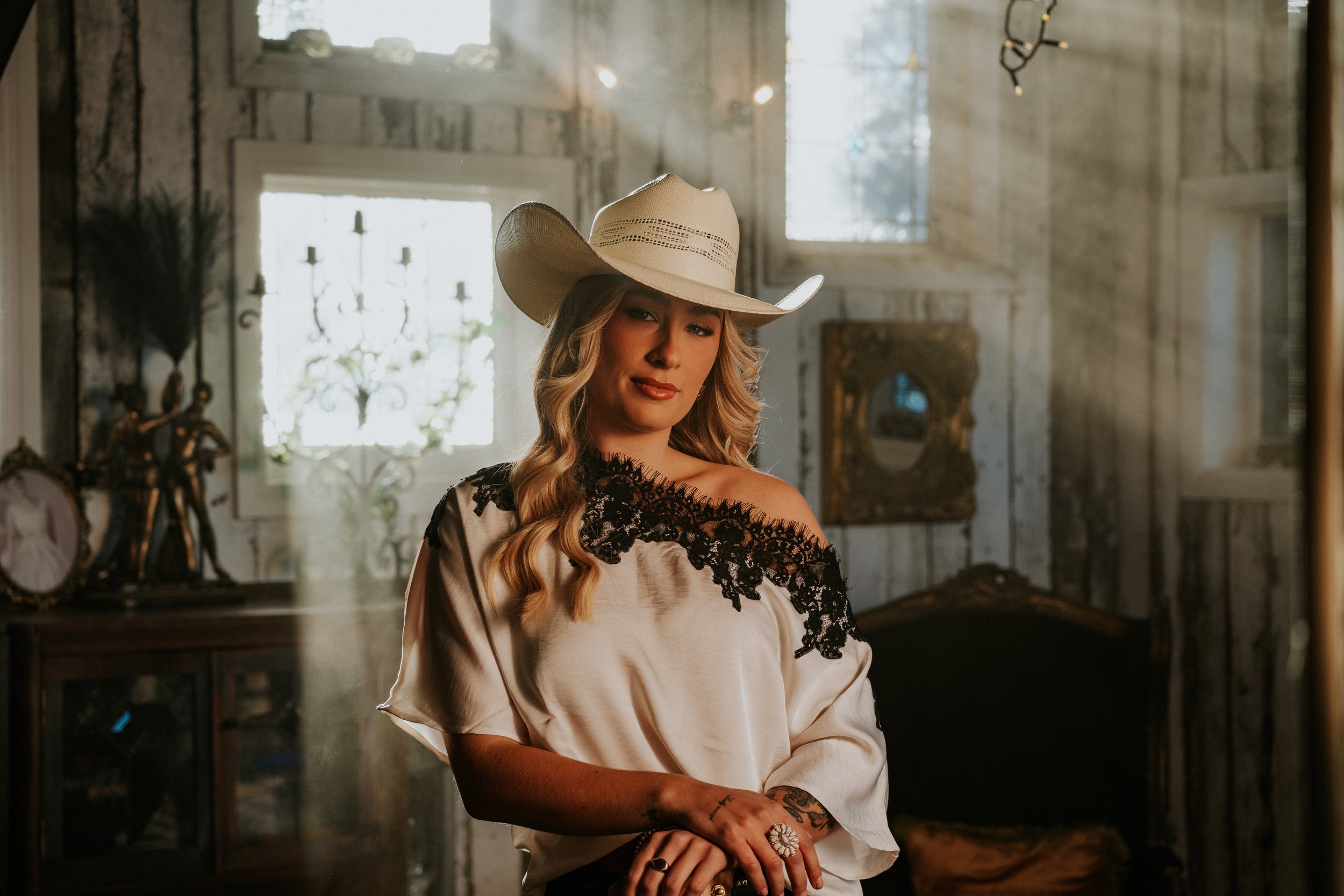 A woman with long, wavy blonde hair wearing a large white cowboy hat and a satin blouse with black lace accents standing in a vintage-style room with antique furniture and decorative objects.